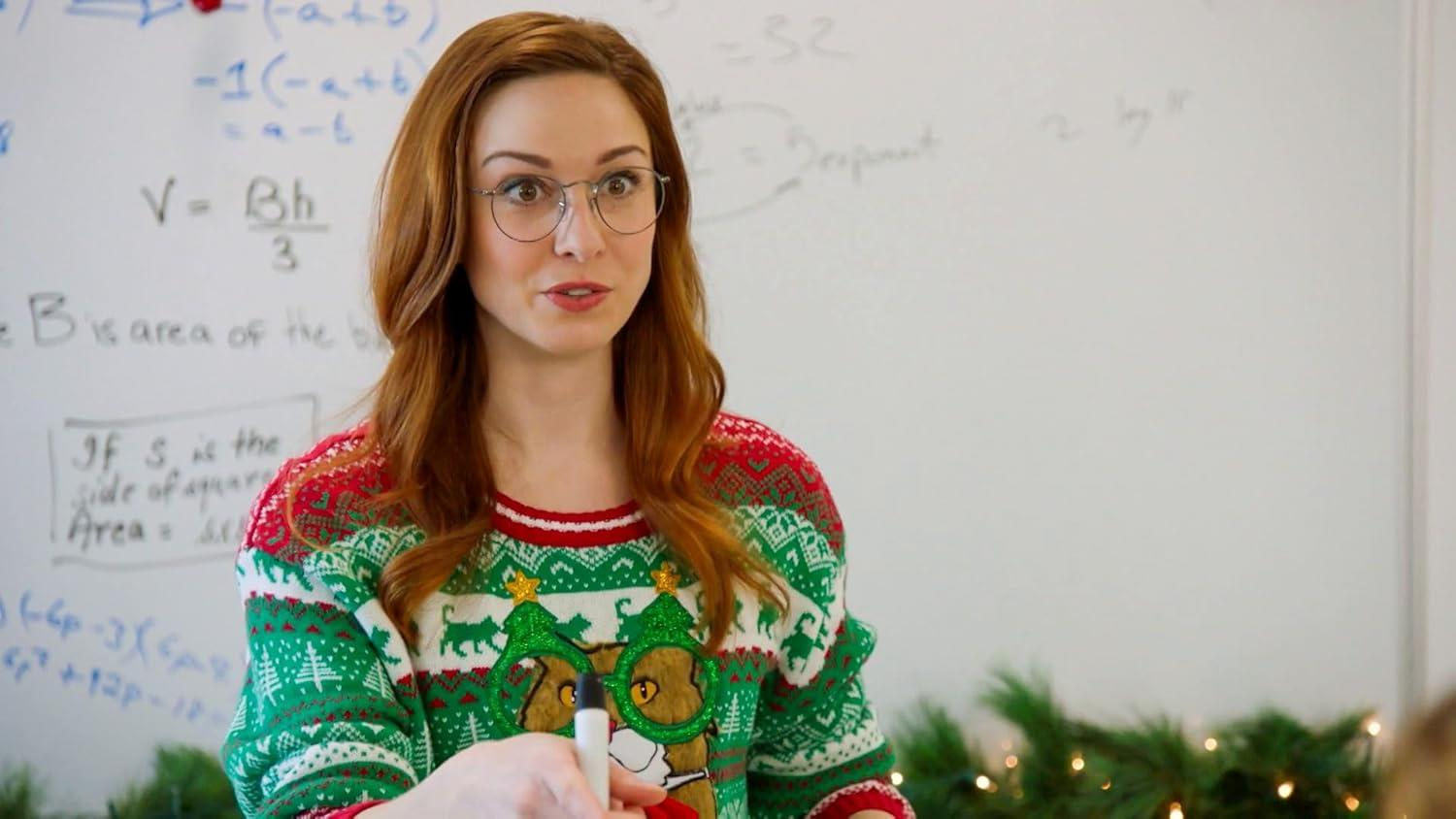 Eine Frau mit Brille und Weihnachtspullover steht vor einer Tafel und hält einen Marker, offenbar beim Unterrichten.