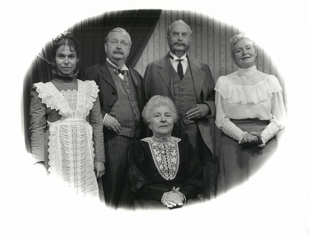 Fünf Personen in Vintage-Kleidung posieren für ein Porträt. Männer in Anzügen, Frauen in Kleidern. Das Bild ist in Schwarzweiß.