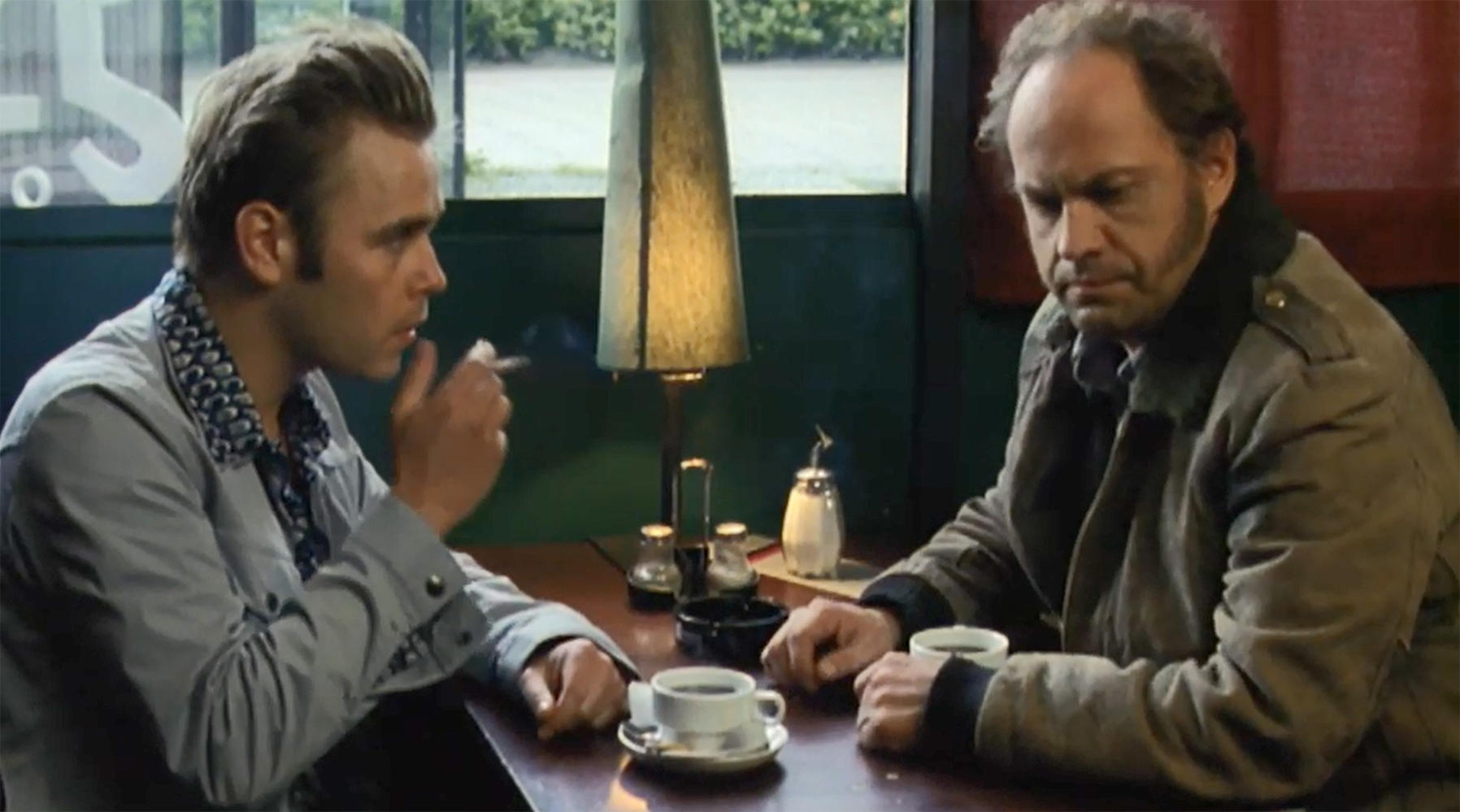 Zwei Männer sitzen an einem Tisch, einer raucht. Kaffeetassen und eine Lampe stehen auf dem Tisch. Dunkler Hintergrund, Retro-Stil.
