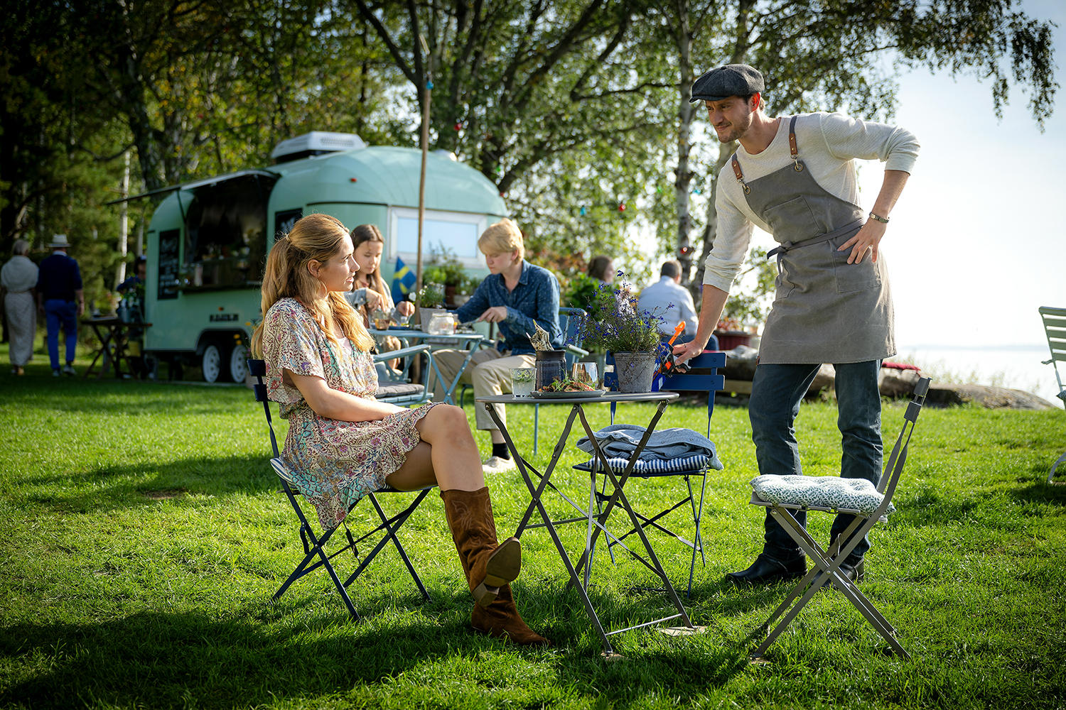 Leute sitzen auf dem Rasen an Tischen. Im Hintergrund ein Foodtruck. Ein Mann mit Schürze steht an einem Tisch.