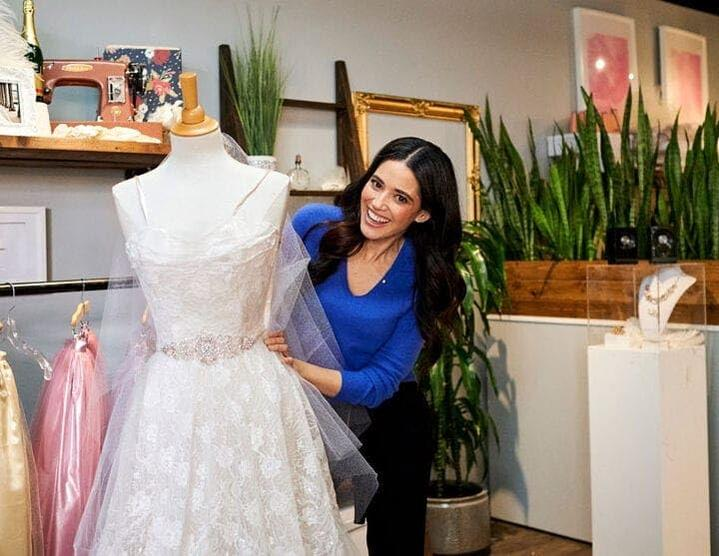 Eine Frau lächelt neben einem Hochzeitskleid auf einer Schaufensterpuppe. Das Kleid ist weißer Spitze, die Frau trägt Blau. Ein Geschäftshintergrund.