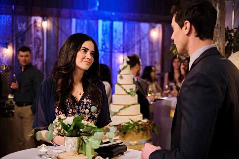 Ein Paar auf einer Hochzeit, Frau blickt Mann an, Kuchen, Blumen und Gäste im Hintergrund. Weiche Beleuchtung und romantisches Ambiente.