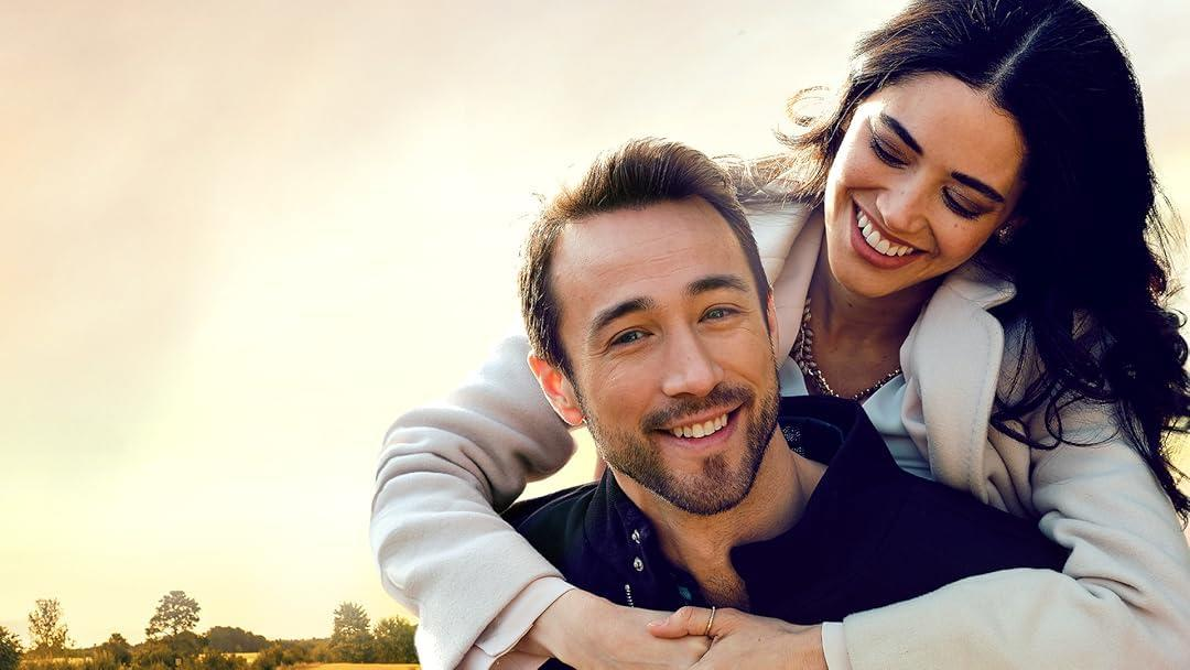 Ein lächelnder Mann wird von einer Frau umarmt. Beide lächeln. Der Hintergrund hat einen gelben Himmel und eine Landschaft.