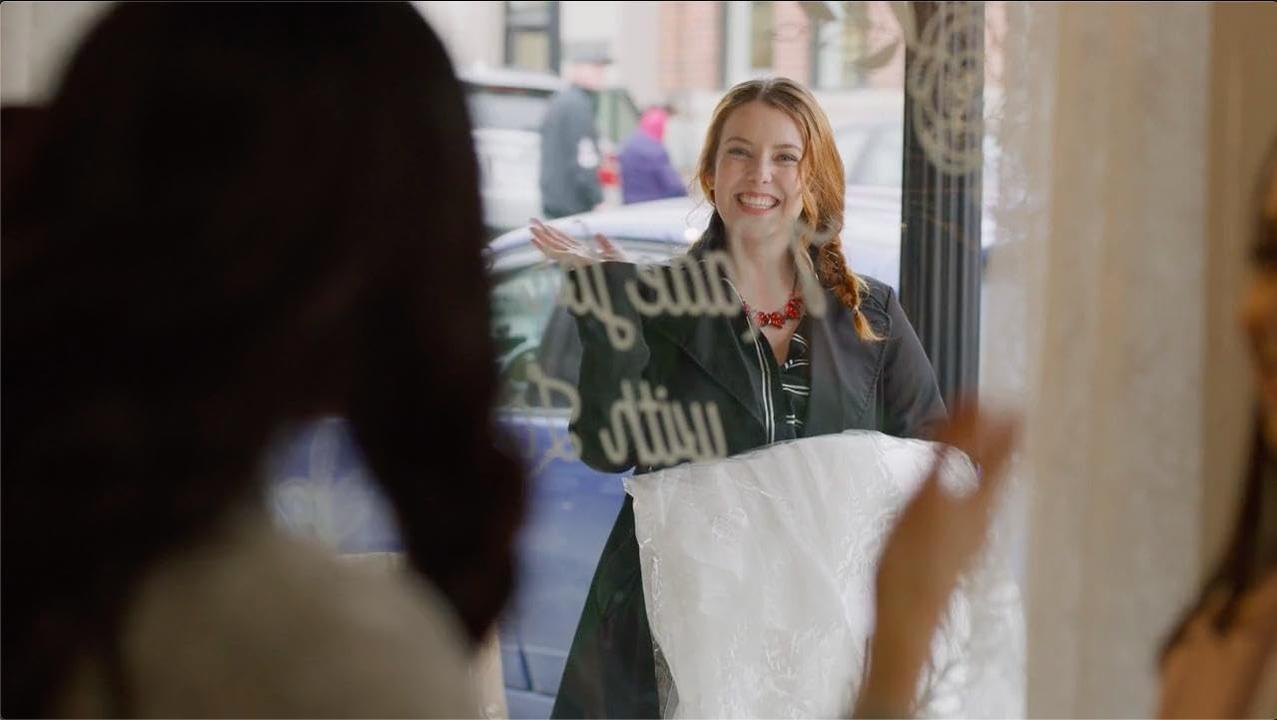 Eine Frau lächelt und hält ein Hochzeitskleid. Sie steht in einem Schaufenster mit Reflexionen anderer. Hinter ihr ist ein Schild.
