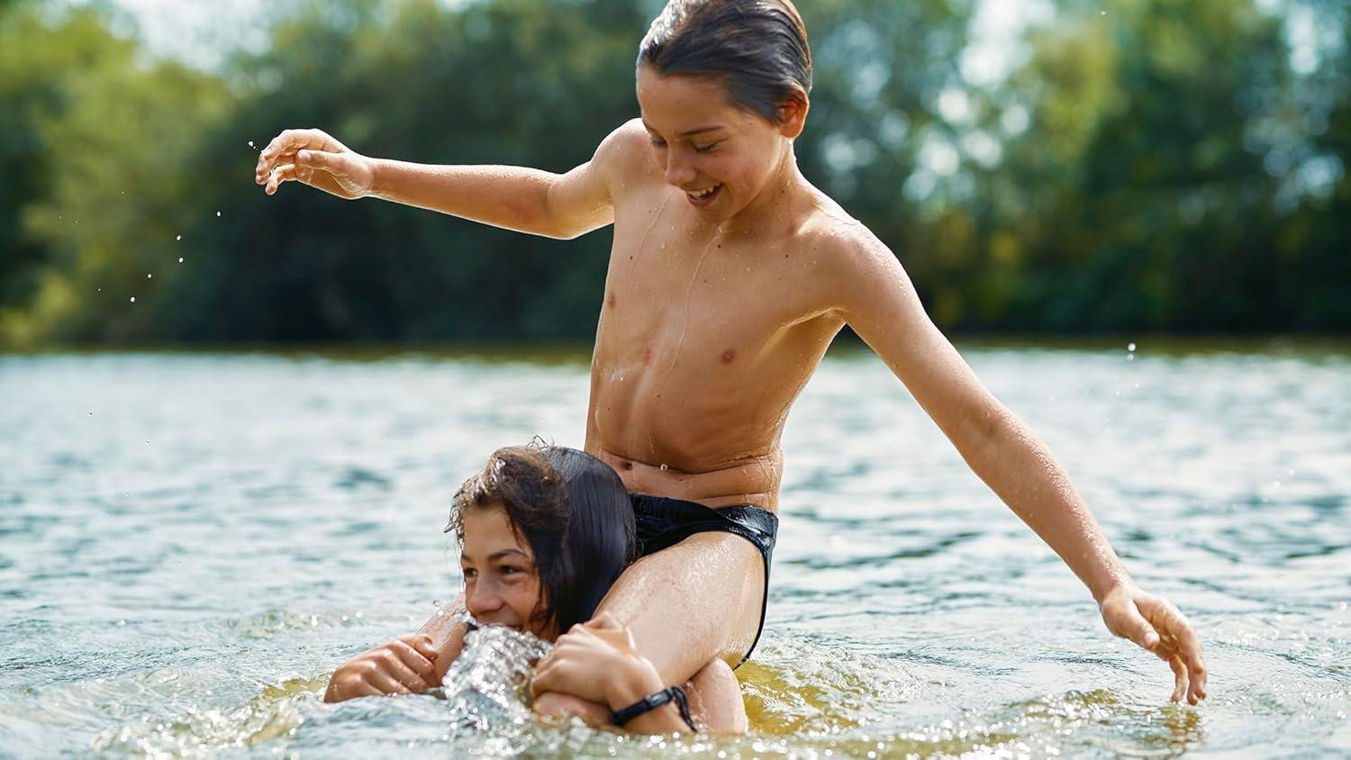 Zwei junge Leute spielen im Wasser. Einer sitzt auf den Schultern des anderen. Sie lächeln und planschen im Wasser.