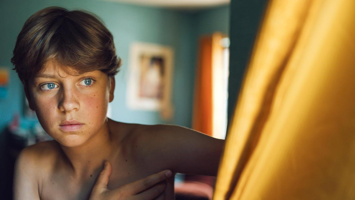 Ein hellhäutiger Junge mit blauen Augen und Sommersprossen blickt nach links. Er ist oberkörperfrei, und rechts ist ein gelber Vorhang.