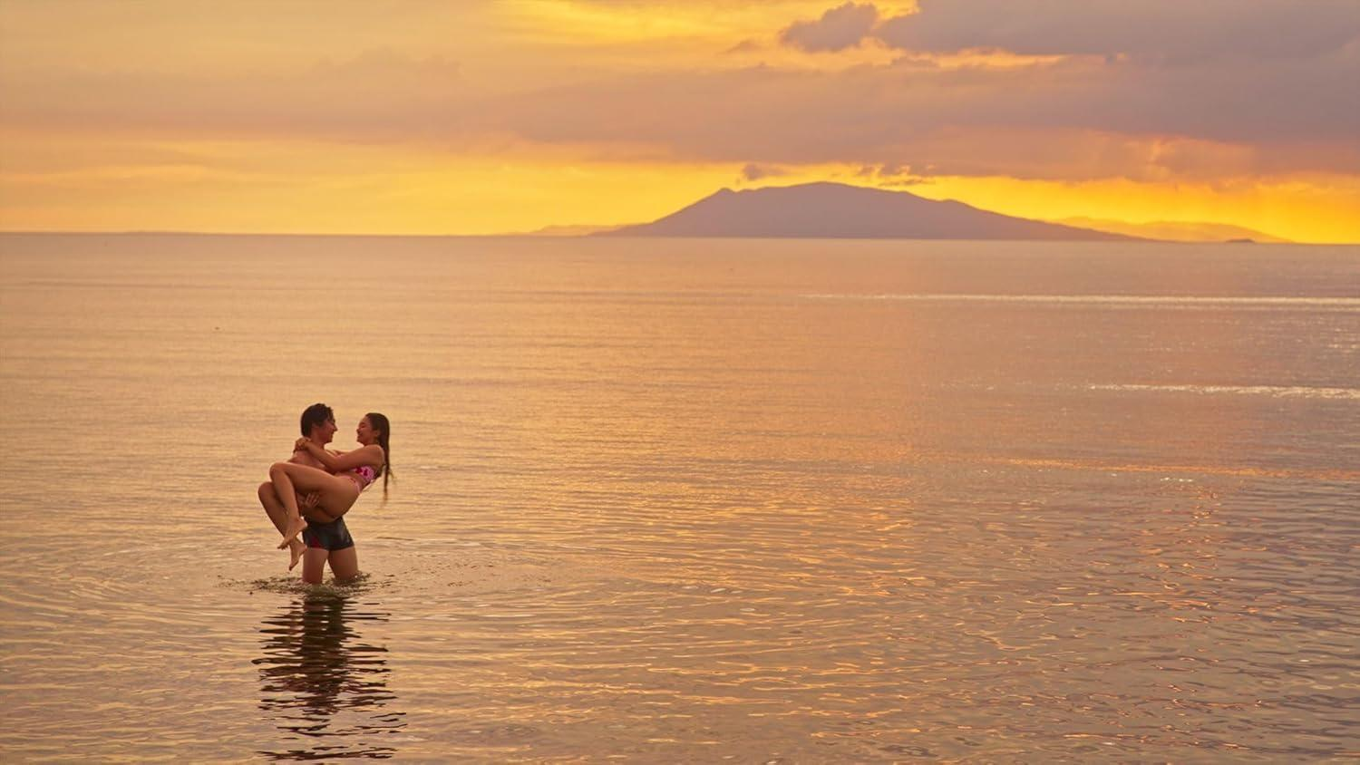Ein Mann trägt eine Frau im Meer bei Sonnenuntergang. Der Himmel ist golden mit einer Bergsilhouette am Horizont.
