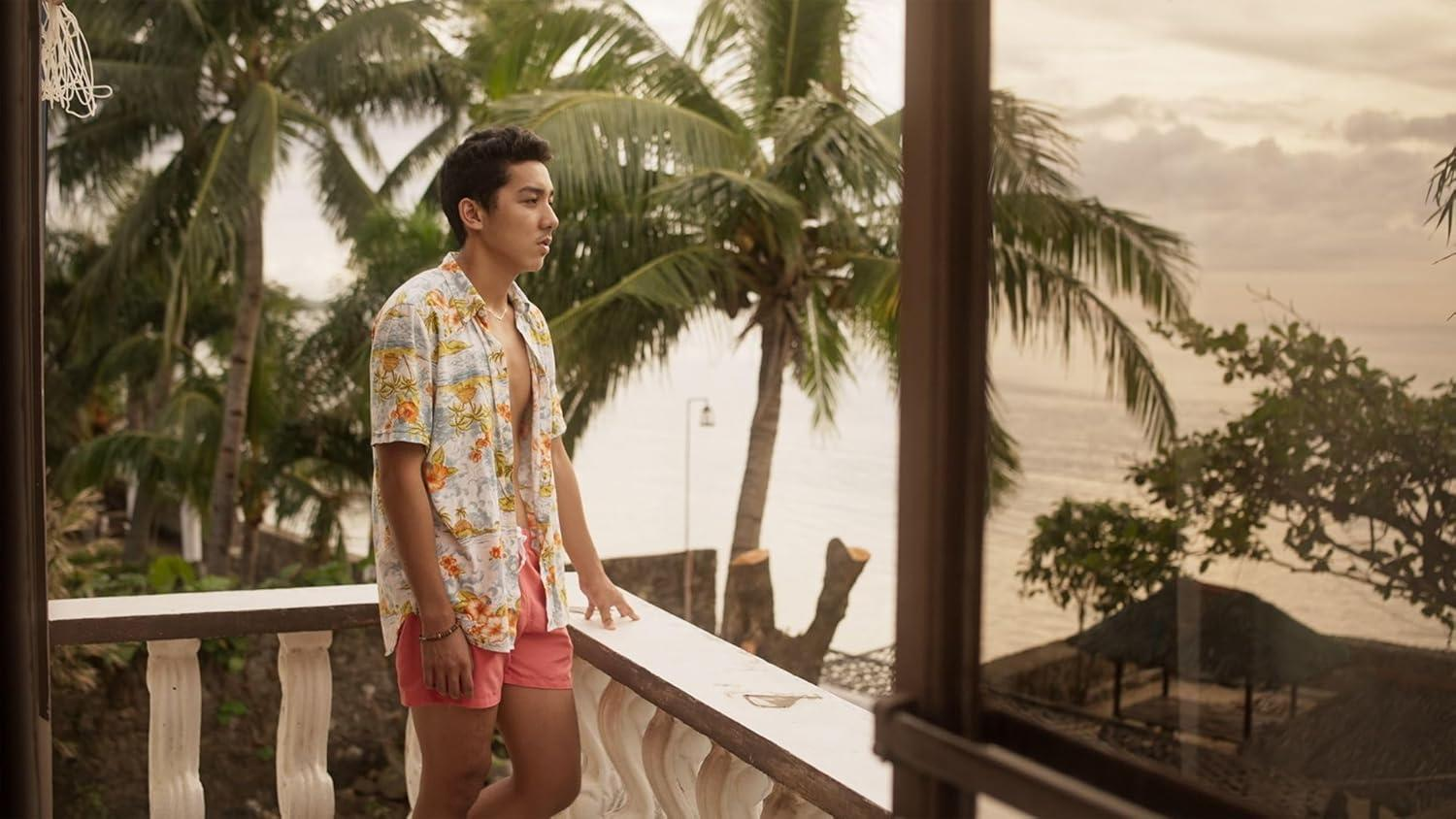Ein Mann in einem Blumenhemd und rosa Shorts steht auf einem Balkon und blickt auf das Meer. Palmen und eine Hütte sind im Hintergrund.