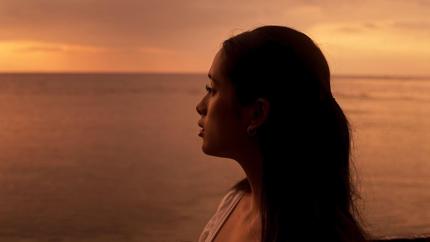 Das Profil einer Frau vor einem Sonnenuntergang über dem Ozean. Der Himmel ist orange, das Wasser ruhig. Sie blickt zum Horizont.