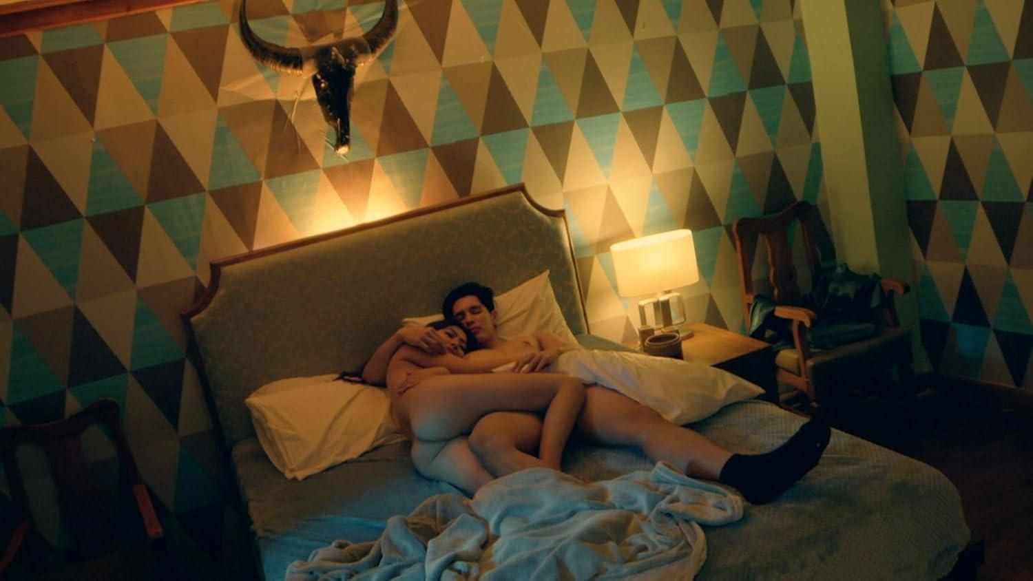 Paar im Bett, umarmt sich. Der Raum hat eine geometrische Tapete, einen Bullenschädel und eine Nachttischlampe. Ein Stuhl ist sichtbar.