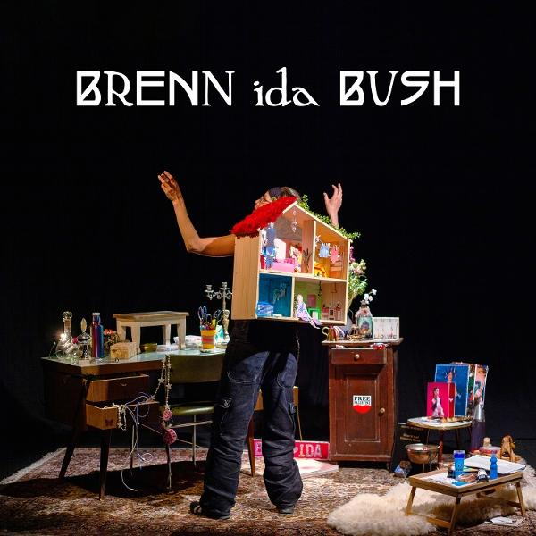 Eine Person hält ein Puppenhaus. Der Hintergrund ist dunkel mit Möbeln und Gegenständen. Oben steht 'BRENN ida BUSH'.