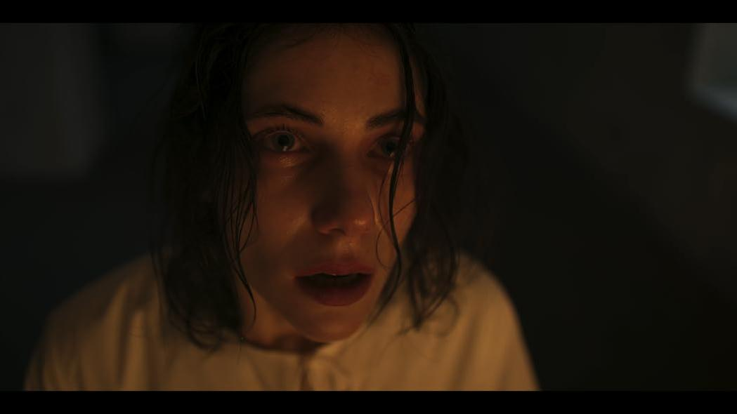 Frau mit aufgerissenen Augen und offenem Mund. Nasses Haar, weißes Hemd und dramatisches Licht deuten auf Angst hin. Dunkler Hintergrund.