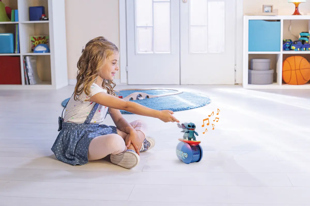 Egy kislány Stitch játékkal játszik, ami egy kék eszköz tetején van. A lány egy szobában ül egy játékvonattal.