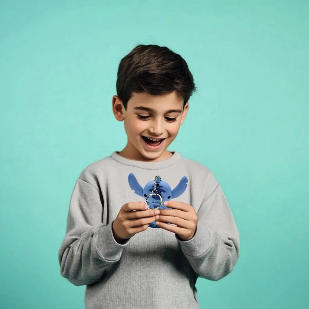 Egy mosolygó fiú kék Stitch kulcstartót tart. Szürke pulóvert visel, türkiz háttér előtt.