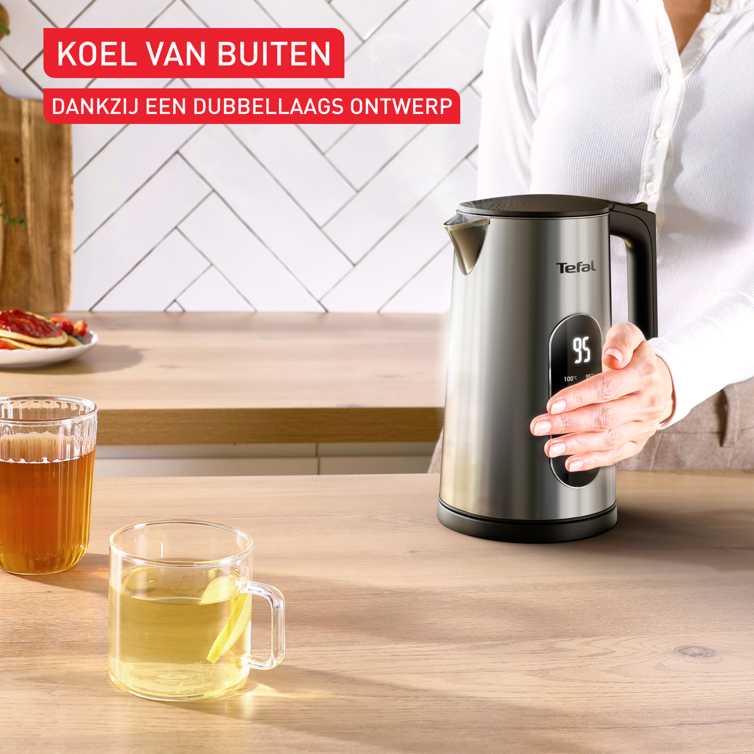 A woman holds a silver kettle with a digital display. Two glasses of tea sit on a wooden countertop.