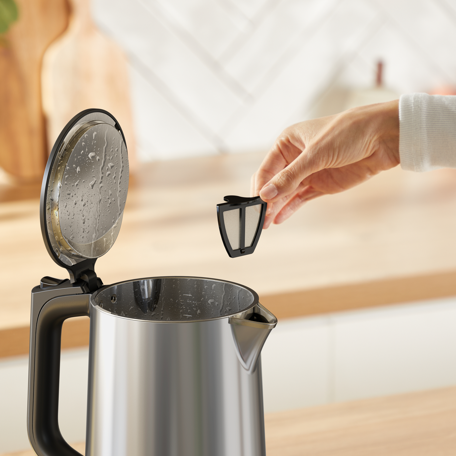 Hand holding a black filter above a stainless steel kettle. The lid is open. Kitchen setting with wood counter.
