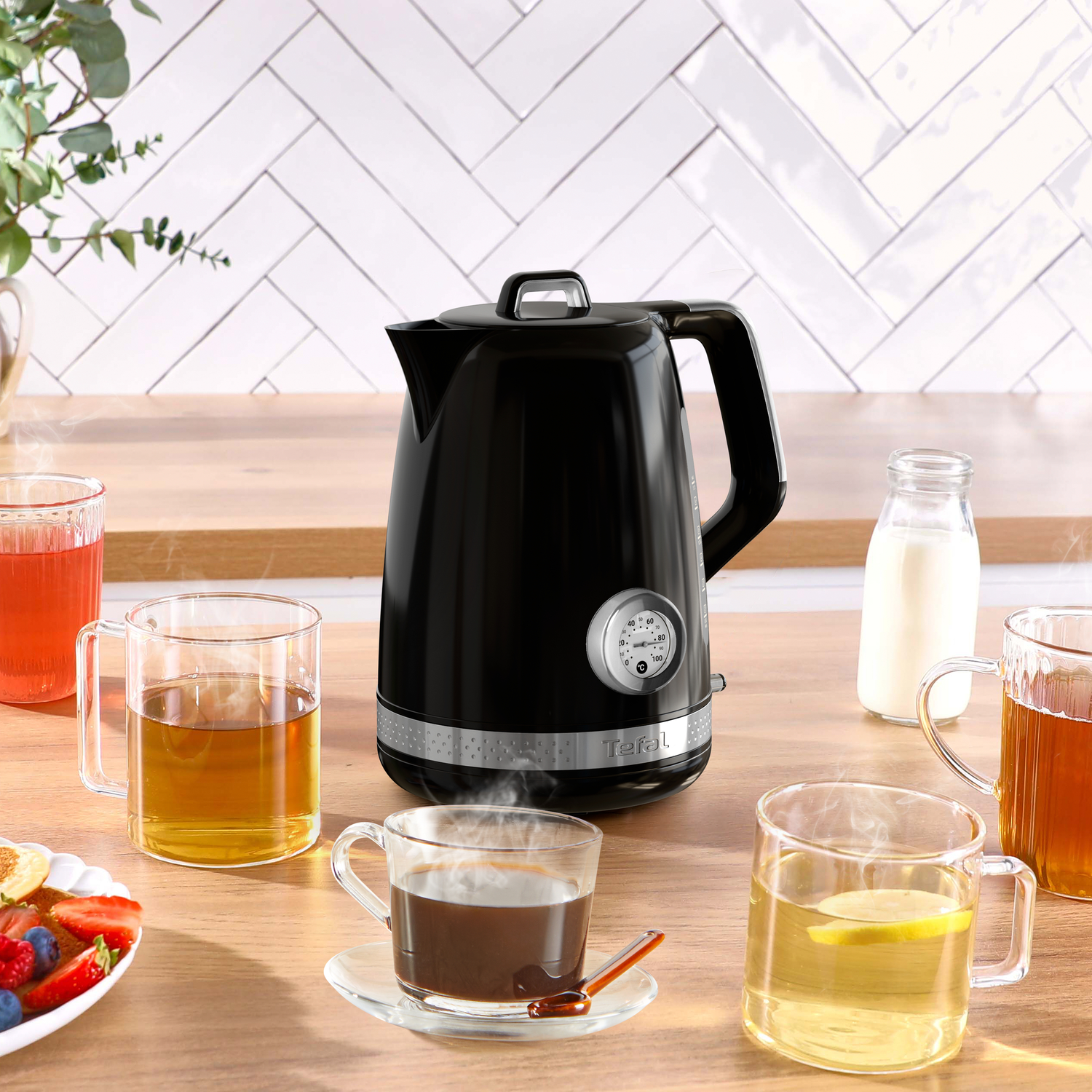 A black electric kettle with tea cups, a milk bottle, and a plate of fruits on a wooden table. White tile background.