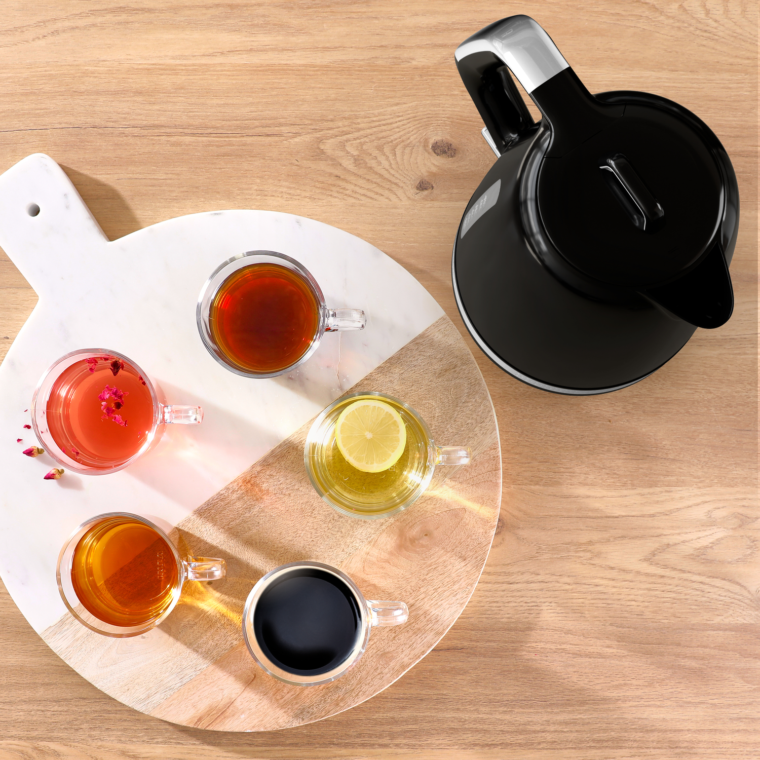 Overhead shot of four tea-filled glass mugs on a wooden board. A black kettle is beside the board on a wooden table.