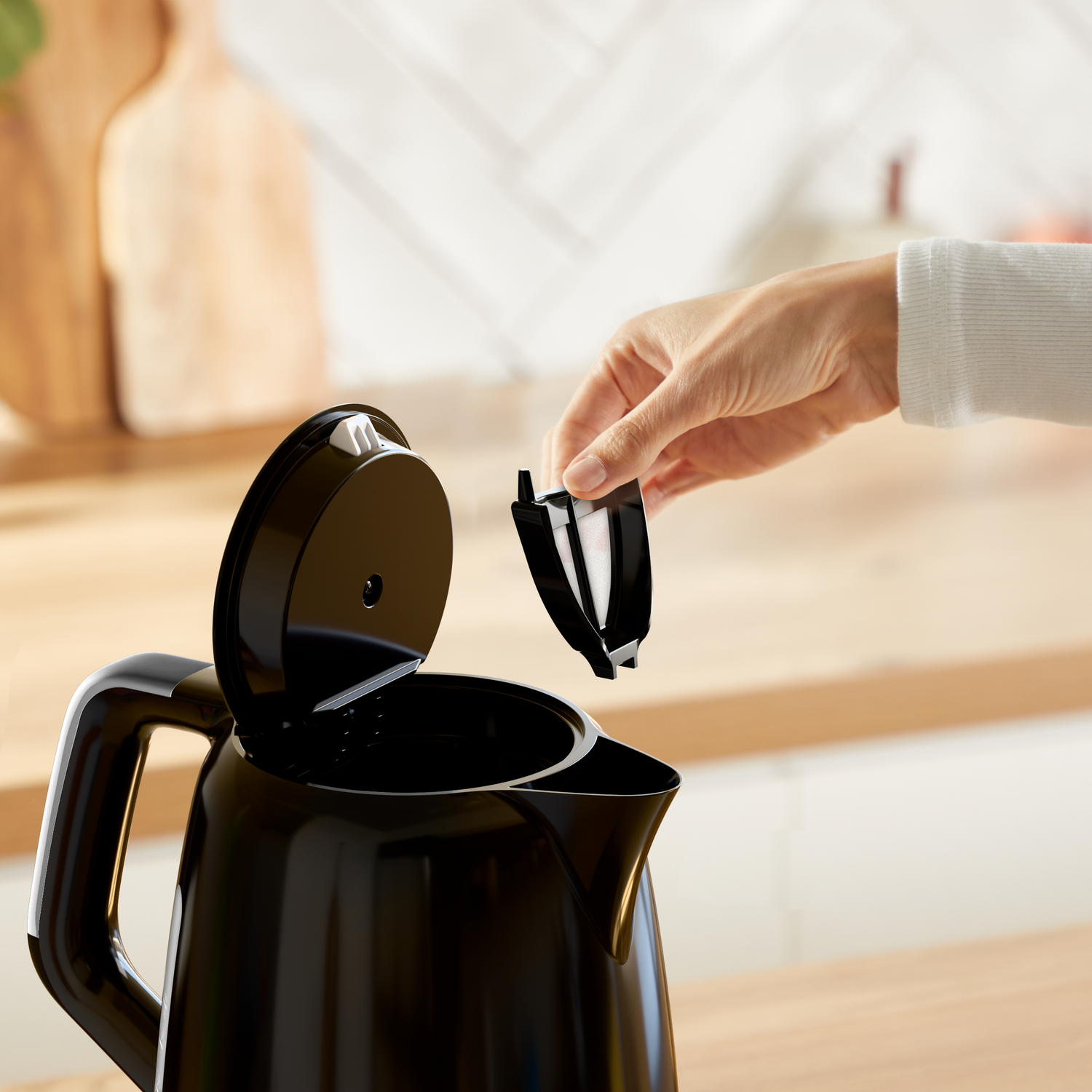 A hand placing a black filter into an open black electric kettle. Kitchen background with wooden counter and white wall.
