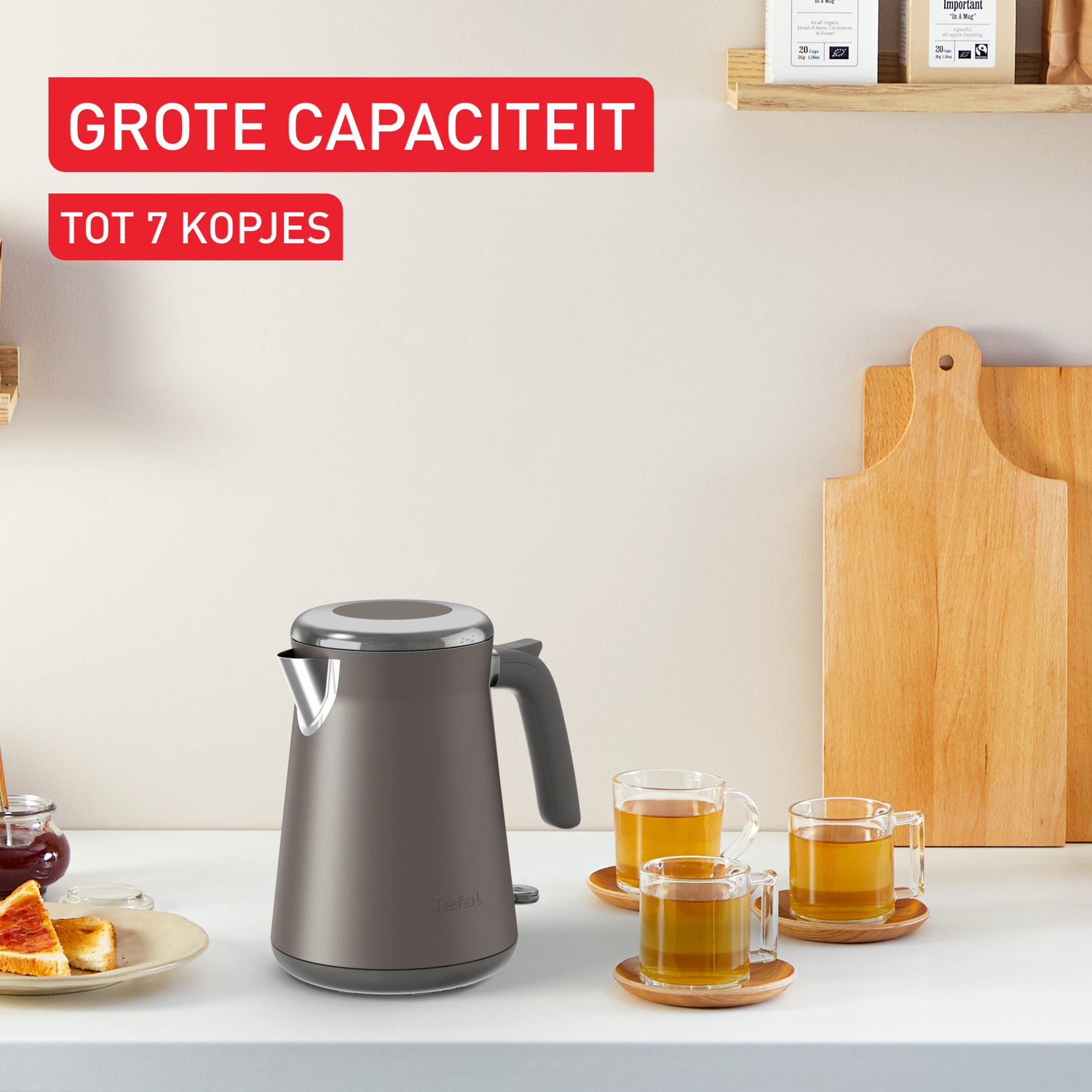 Een grijze waterkoker, drie theekopjes en toast met jam op een wit oppervlak. De achtergrond bevat houten snijplanken.