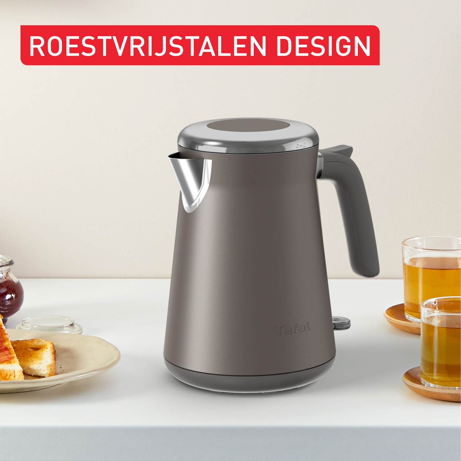 Een grijze waterkoker met een zwart handvat staat op een witte tafel. Toast en jam staan links. Twee glazen thee staan rechts.