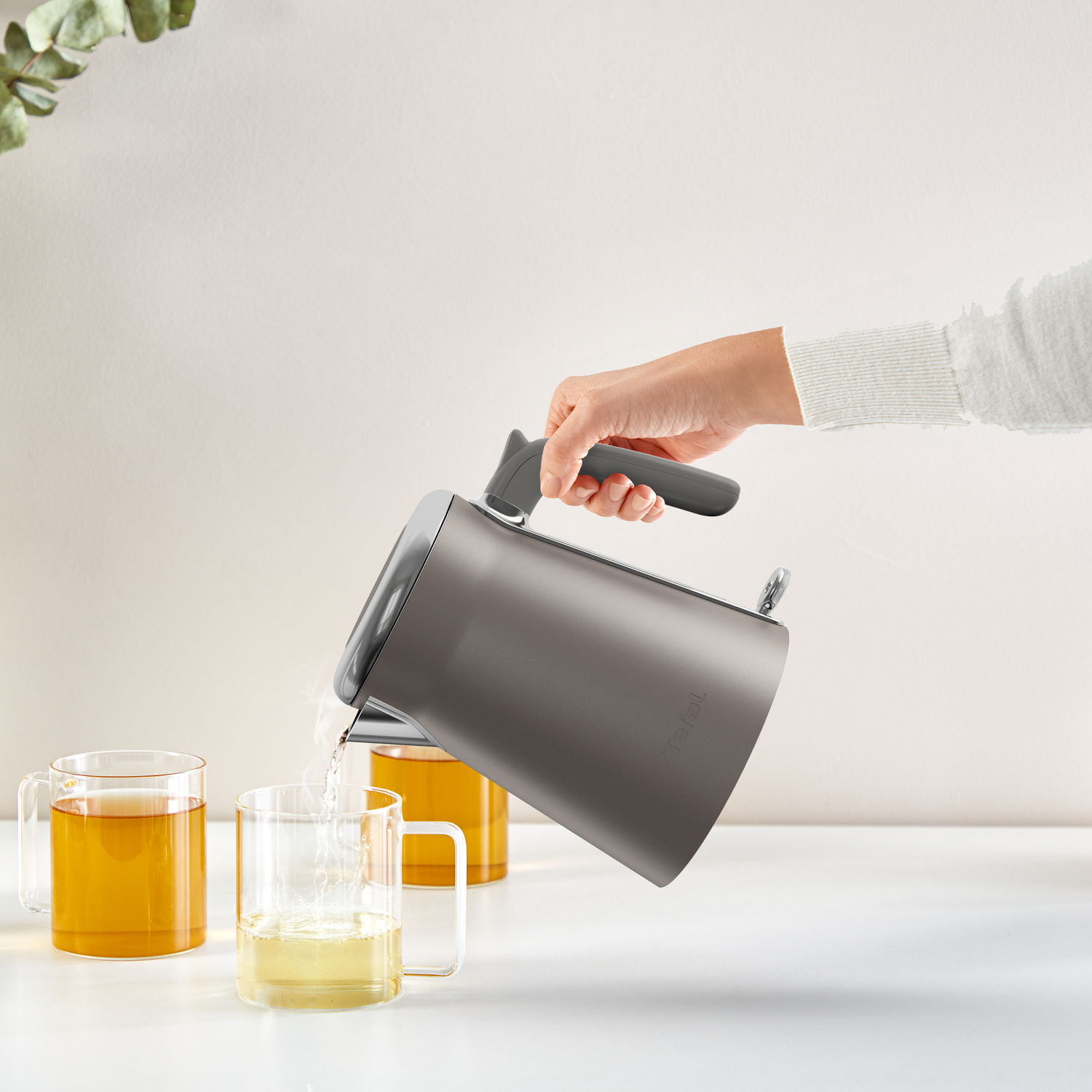 Een hand giet water uit een grijze waterkoker in een glazen mok. Twee andere mokken met thee staan op tafel en een plant is zichtbaar.