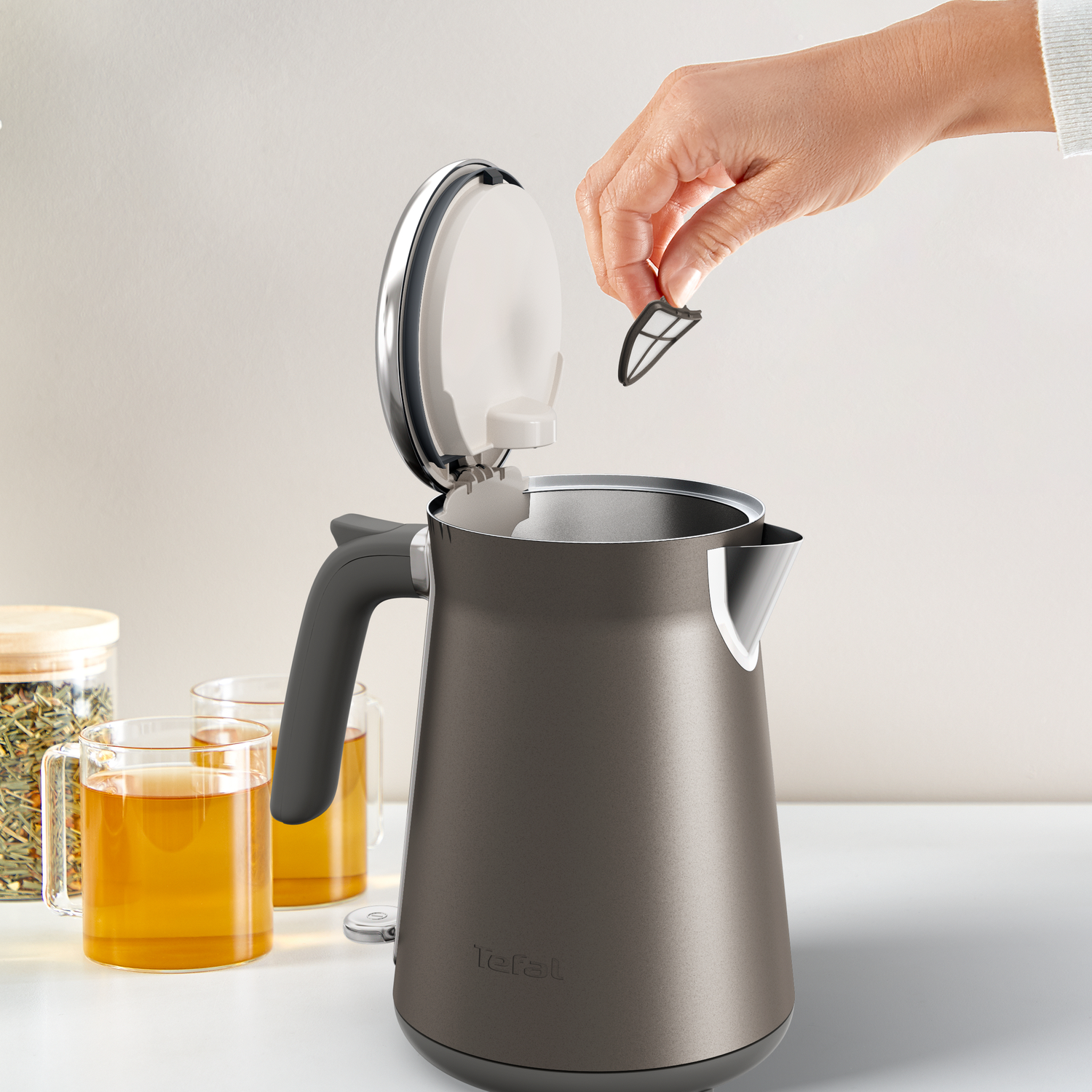 Een hand plaatst een theefilter in een open grijze waterkoker. Theekopjes en een theepot staan op de witte tafel.