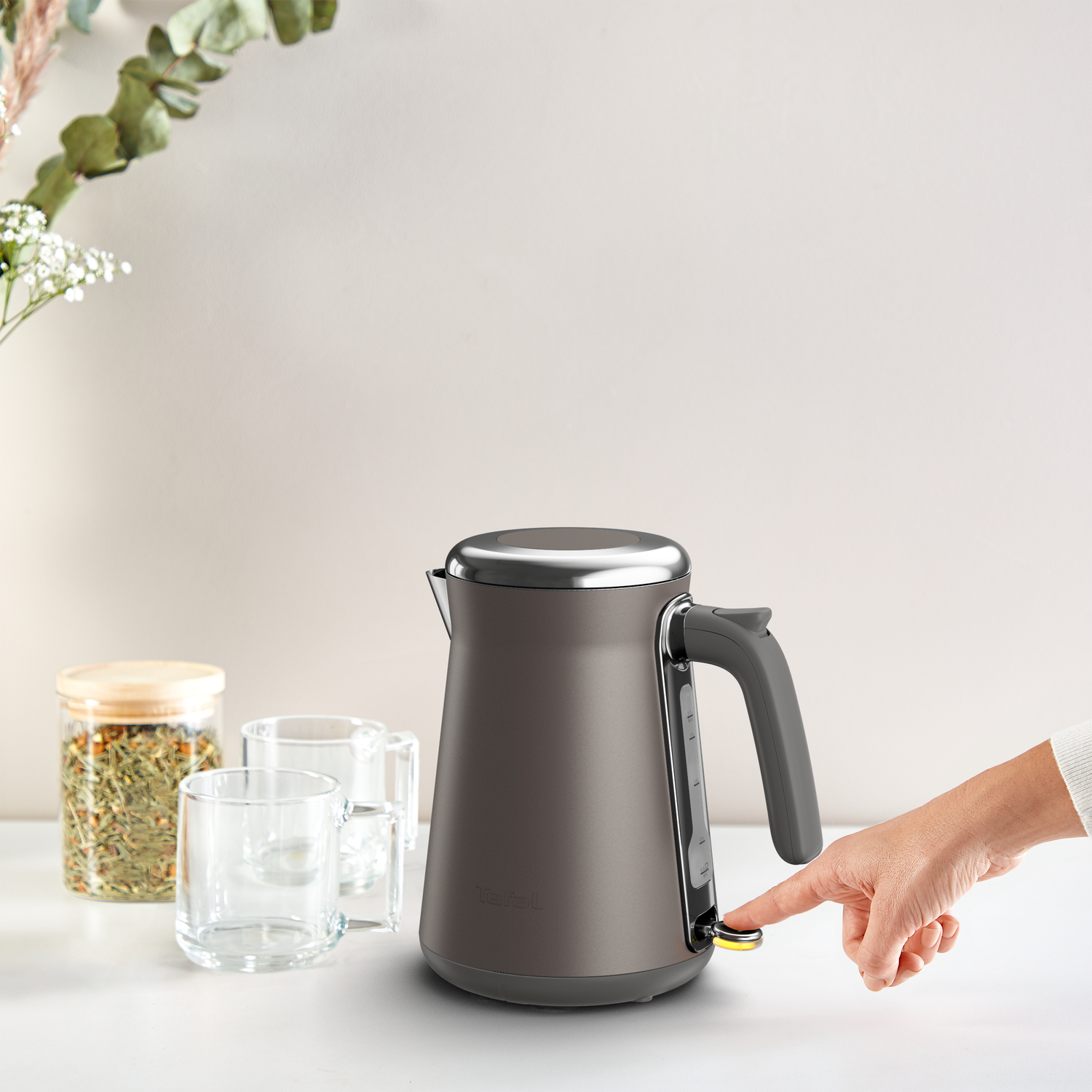 Een hand drukt op een knop op een grijze waterkoker. Twee glazen, een pot thee en groen staan ook op tafel.