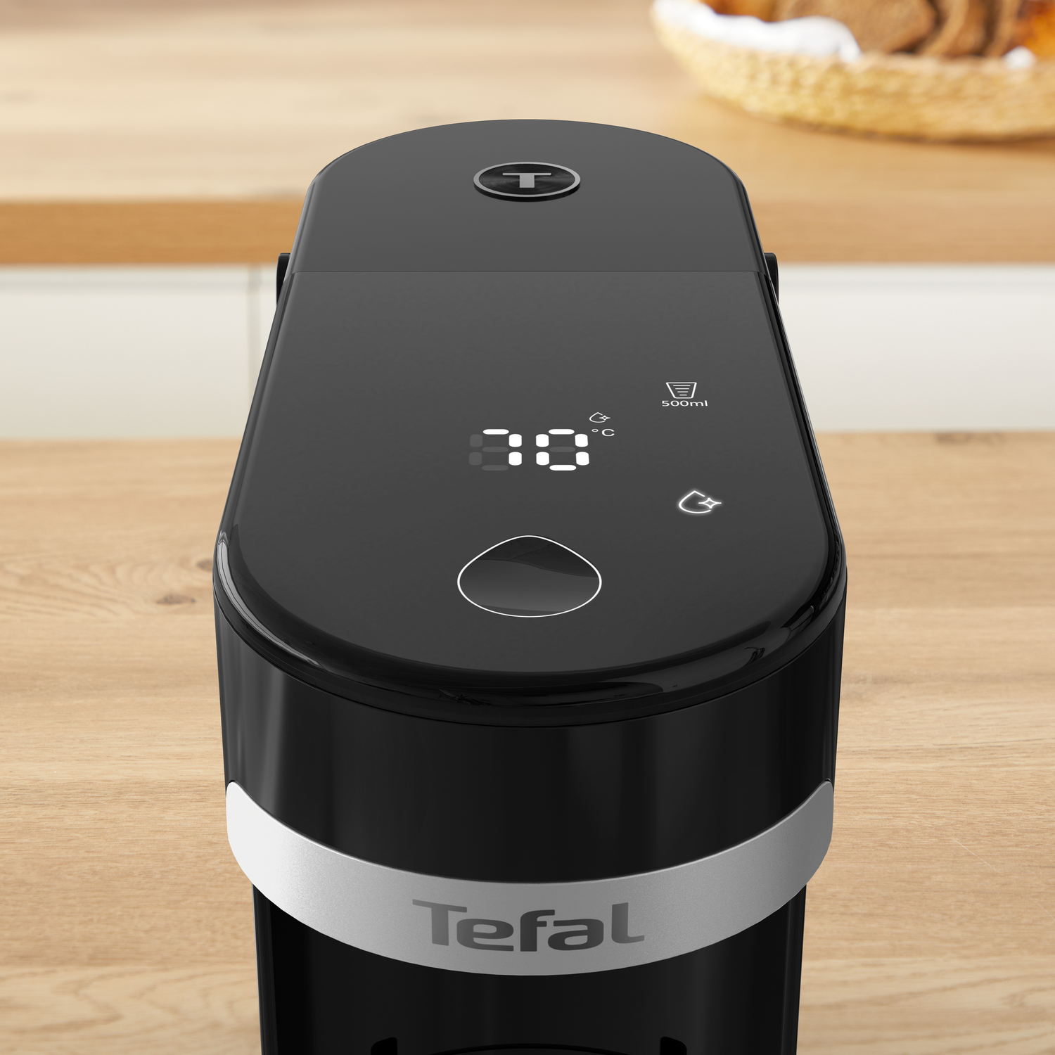 Appareil Tefal noir sur une surface en bois. Il possède un affichage numérique de la température. Un panier et une surface en bois sont en arrière-plan.