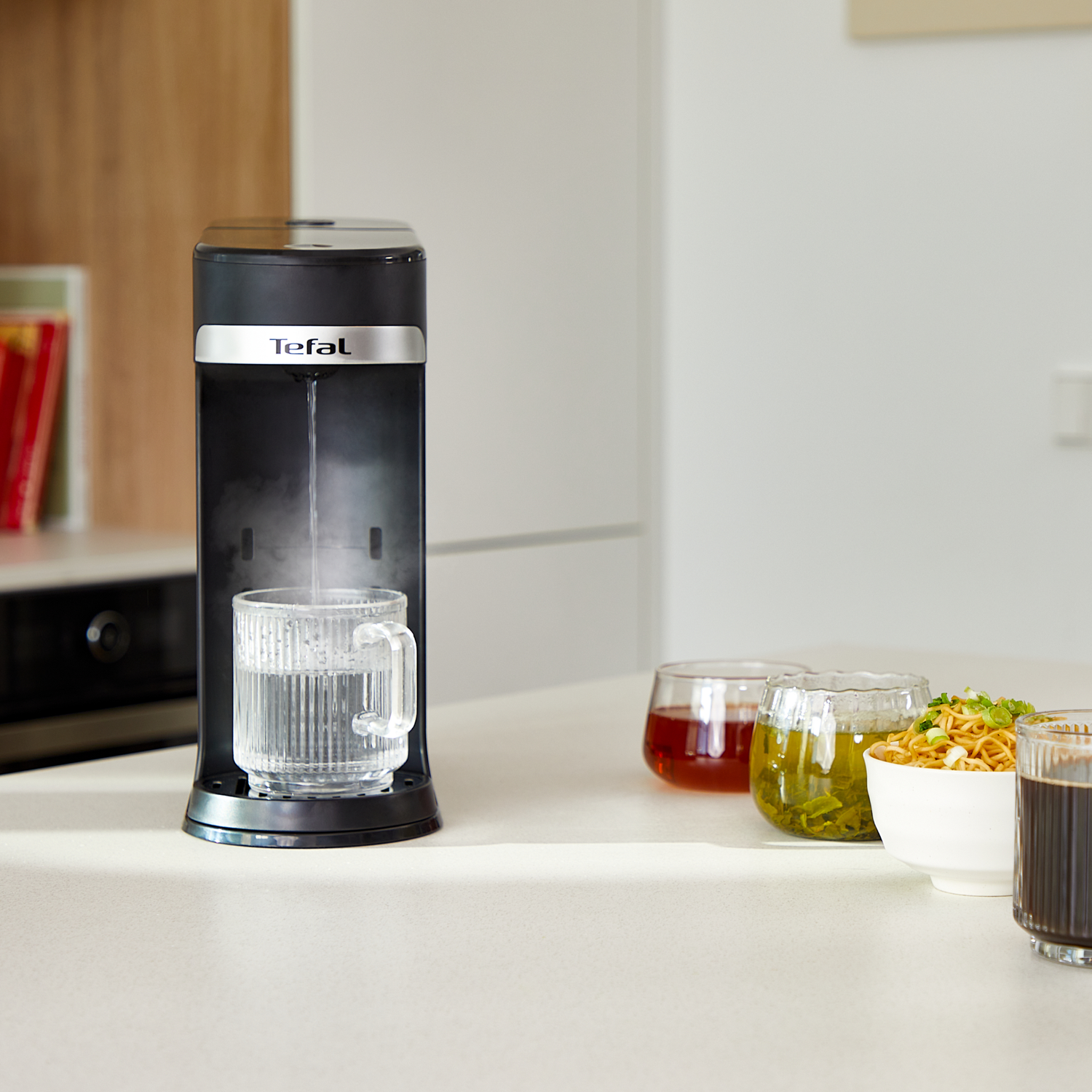 Un distributeur d'eau noir remplit un verre transparent. Des bols de nouilles et de boissons sont sur la table. L'arrière-plan est une cuisine.