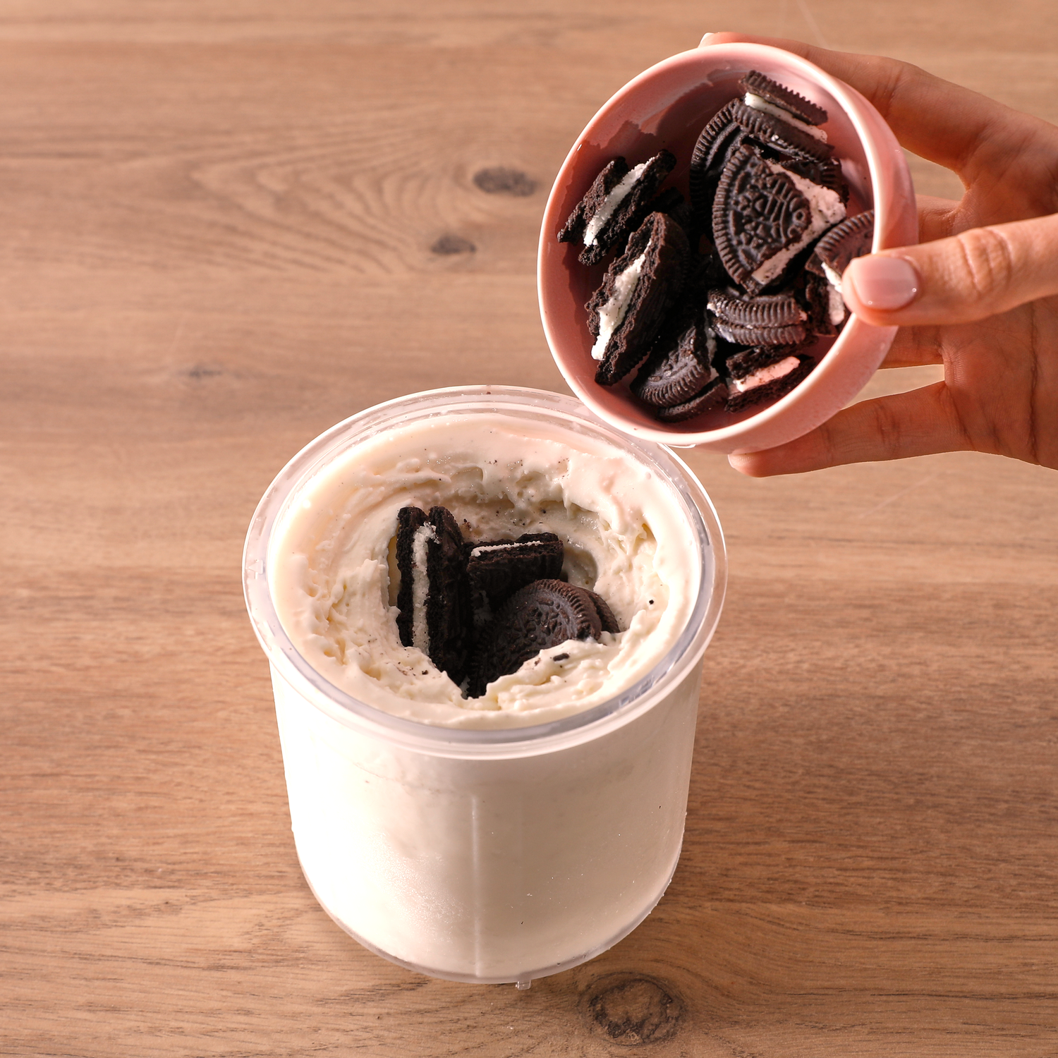 Een hand giet Oreo-koekjes uit een roze kom in een bak ijs. De achtergrond is een houten oppervlak.