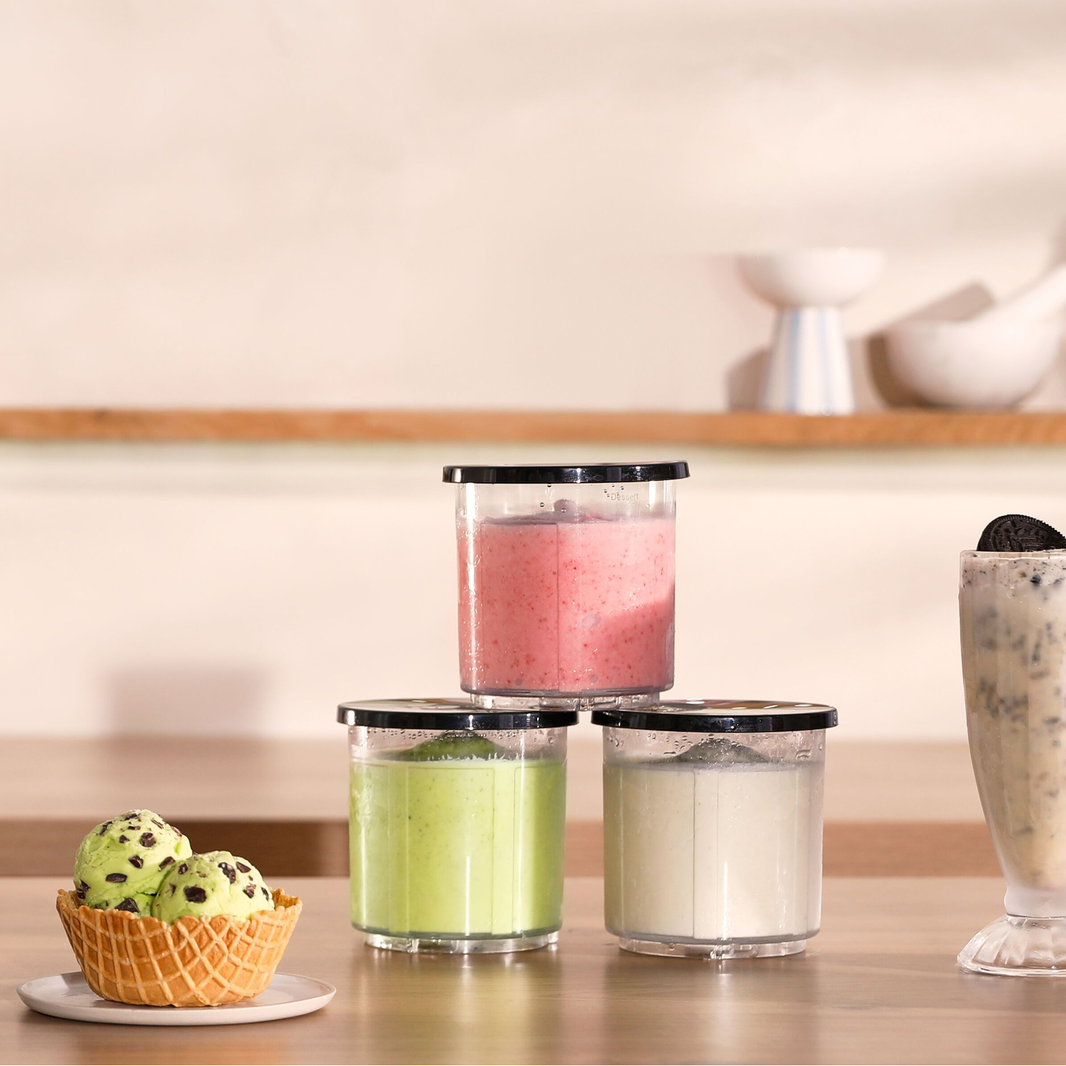 Een tafel toont drie smoothies in doorzichtige containers, gerangschikt in een piramide. Een wafelbeker met ijs en een Oreo-shake zijn ook zichtbaar.