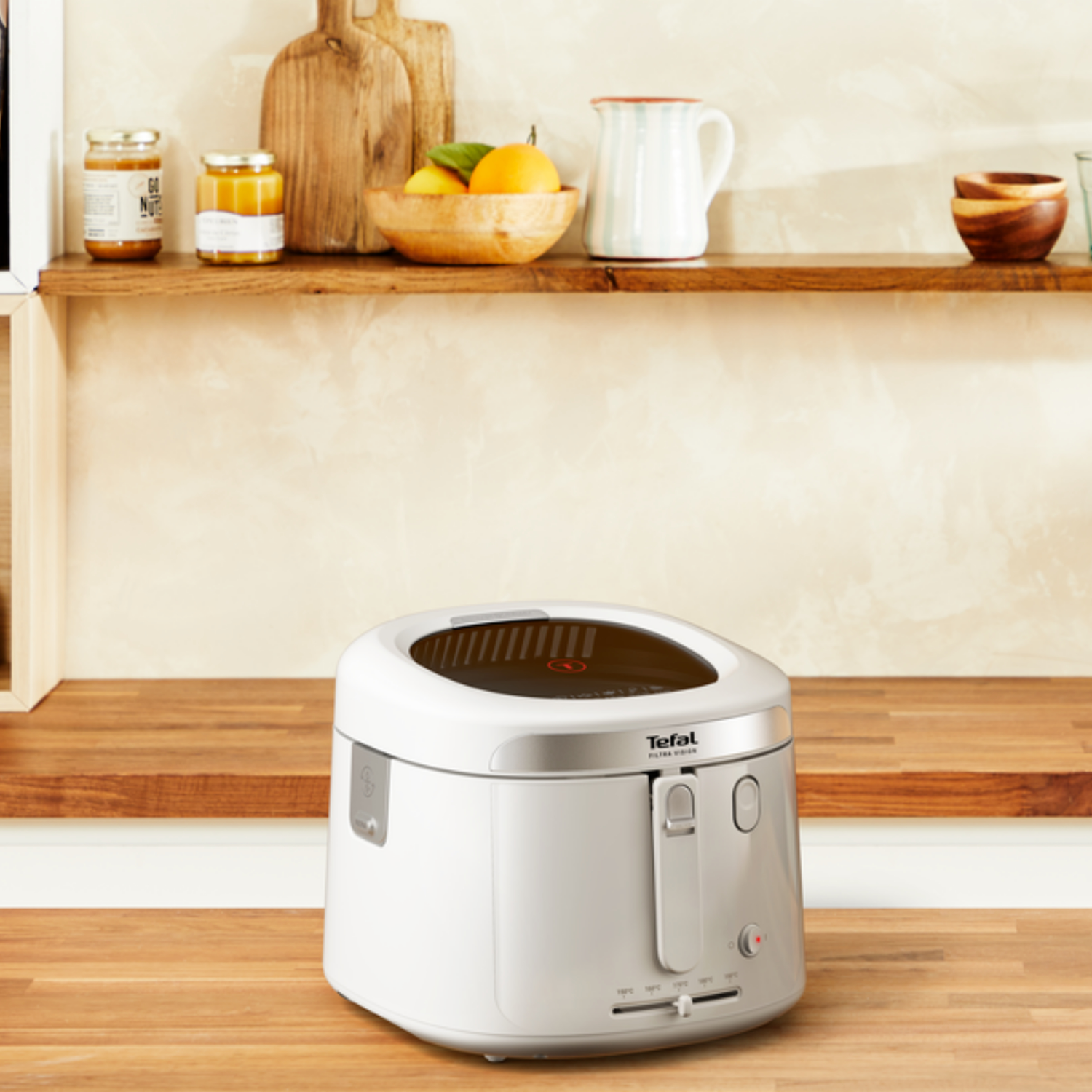 A white Tefal deep fryer sits on a wooden counter. Shelves above hold jars, cutting boards, a bowl of oranges, and a pitcher.