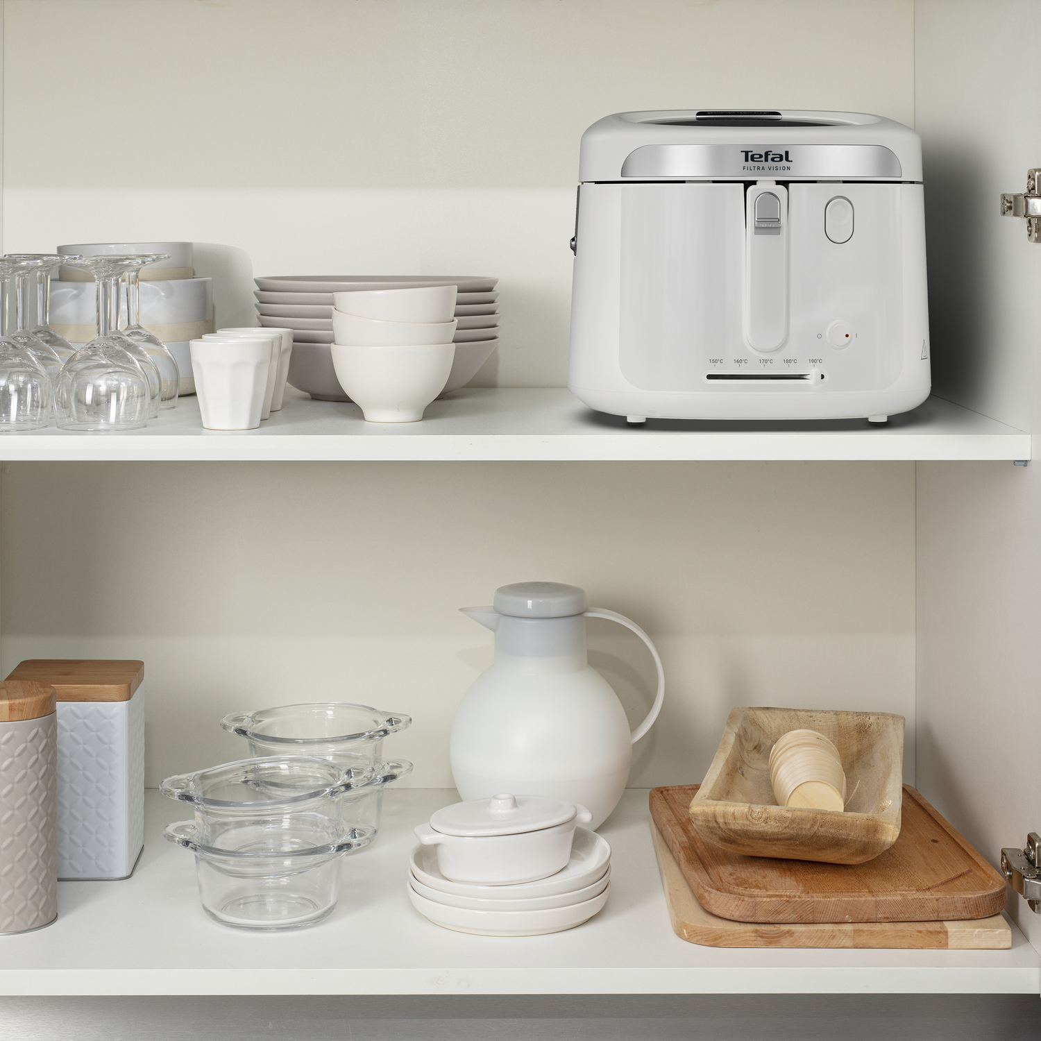 A white kitchen cabinet with various items. It includes a deep fryer, plates, cups, glasses, a pitcher, and wooden cutting boards.
