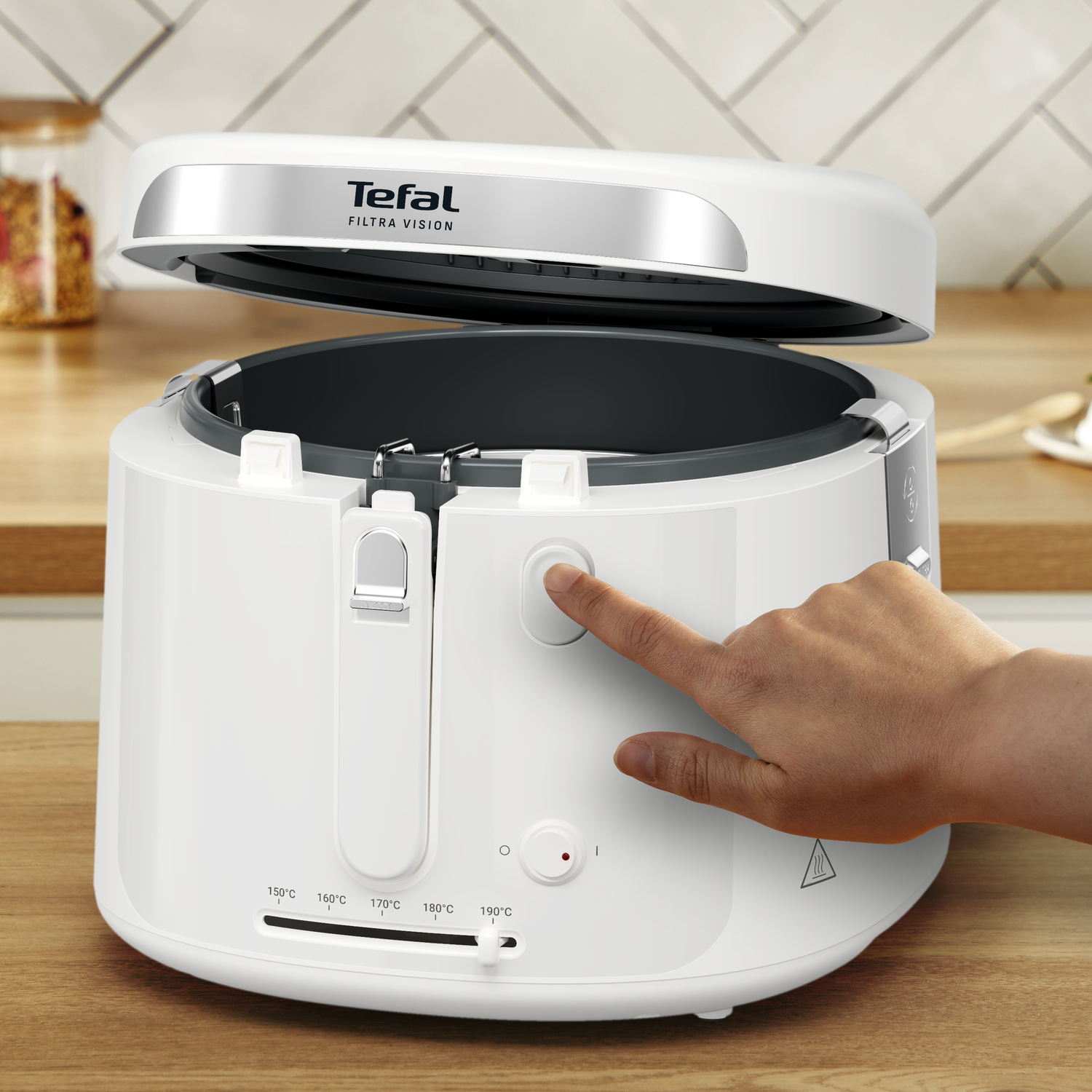 A hand presses the button on a white Tefal deep fryer. The lid is open. Temperature settings are visible.
