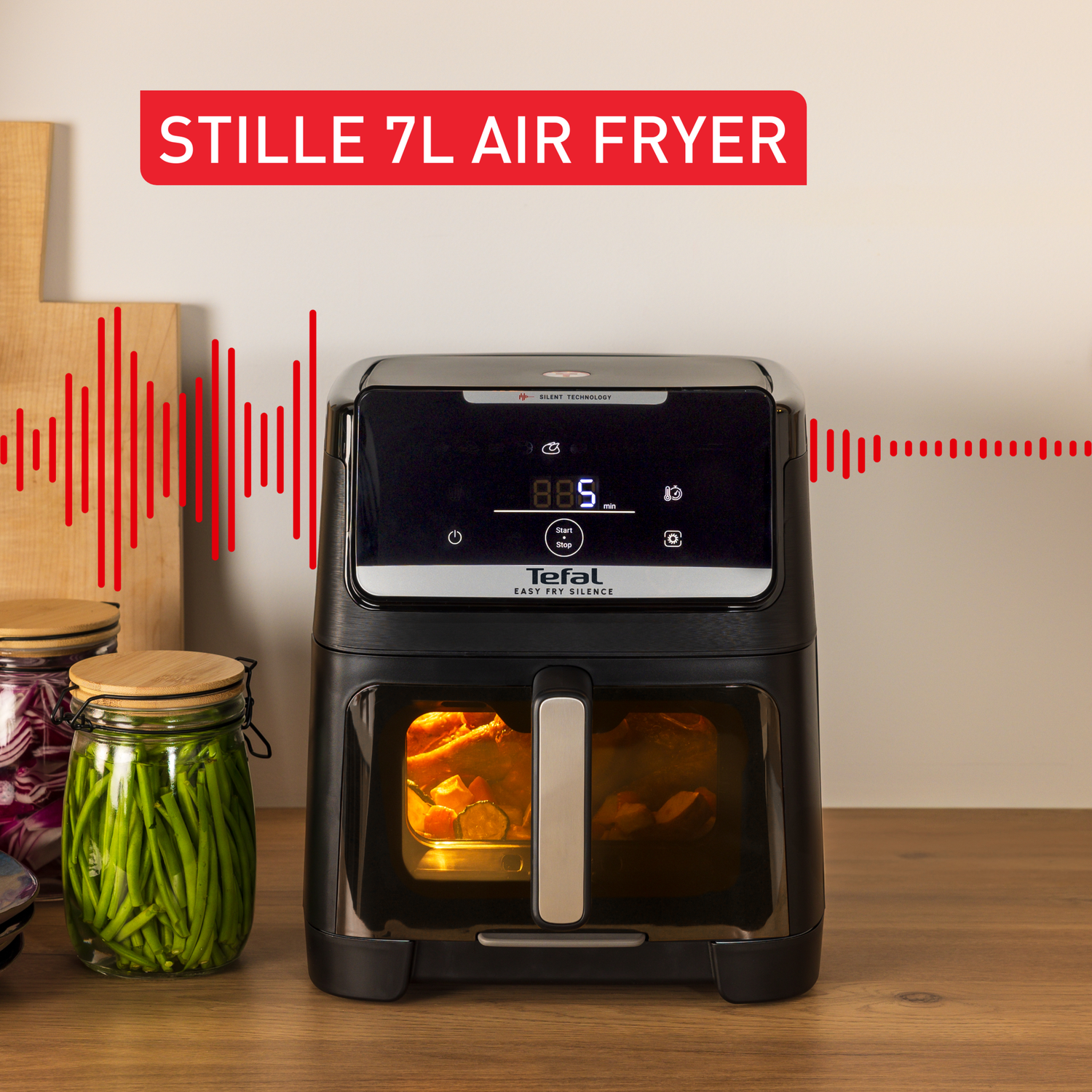 Black air fryer on a wooden surface, displaying the time. Jars of vegetables are on the left. Red text at the top says 'STILLE 7L AIR FRYER'.