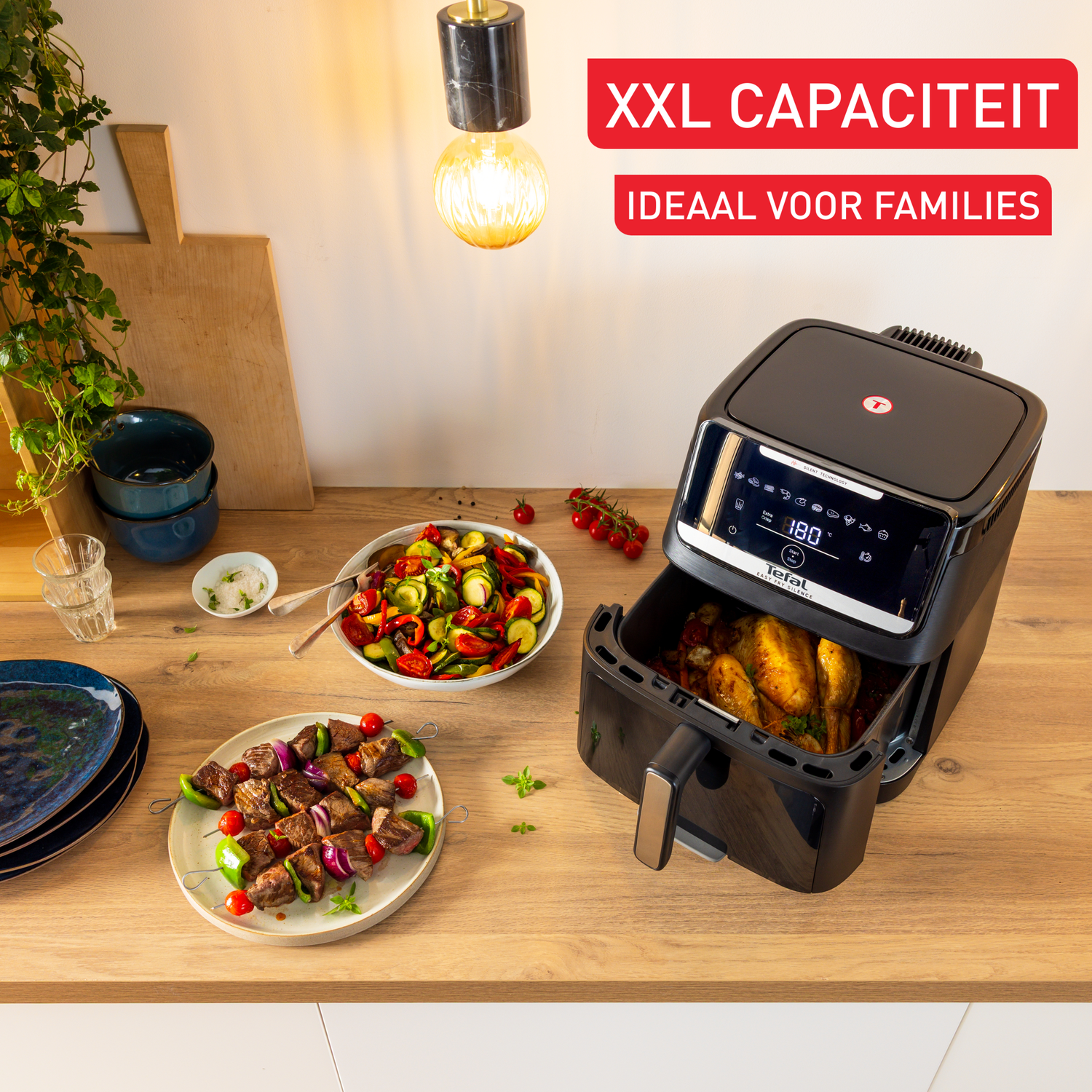 An air fryer with a chicken inside. There are plates of food like vegetables and kebabs. The text above says XXL CAPACITEIT.