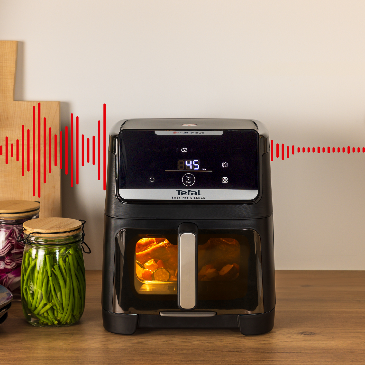 Black air fryer on a wooden surface. Vegetables in jars on the left. Red sound waves on the top left and right.
