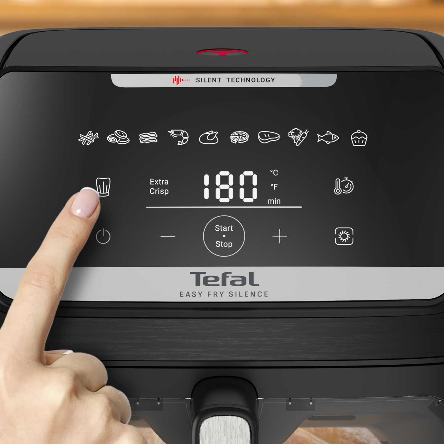 A hand presses the power button on a black air fryer. The display shows 180 degrees. Icons for different foods are visible.