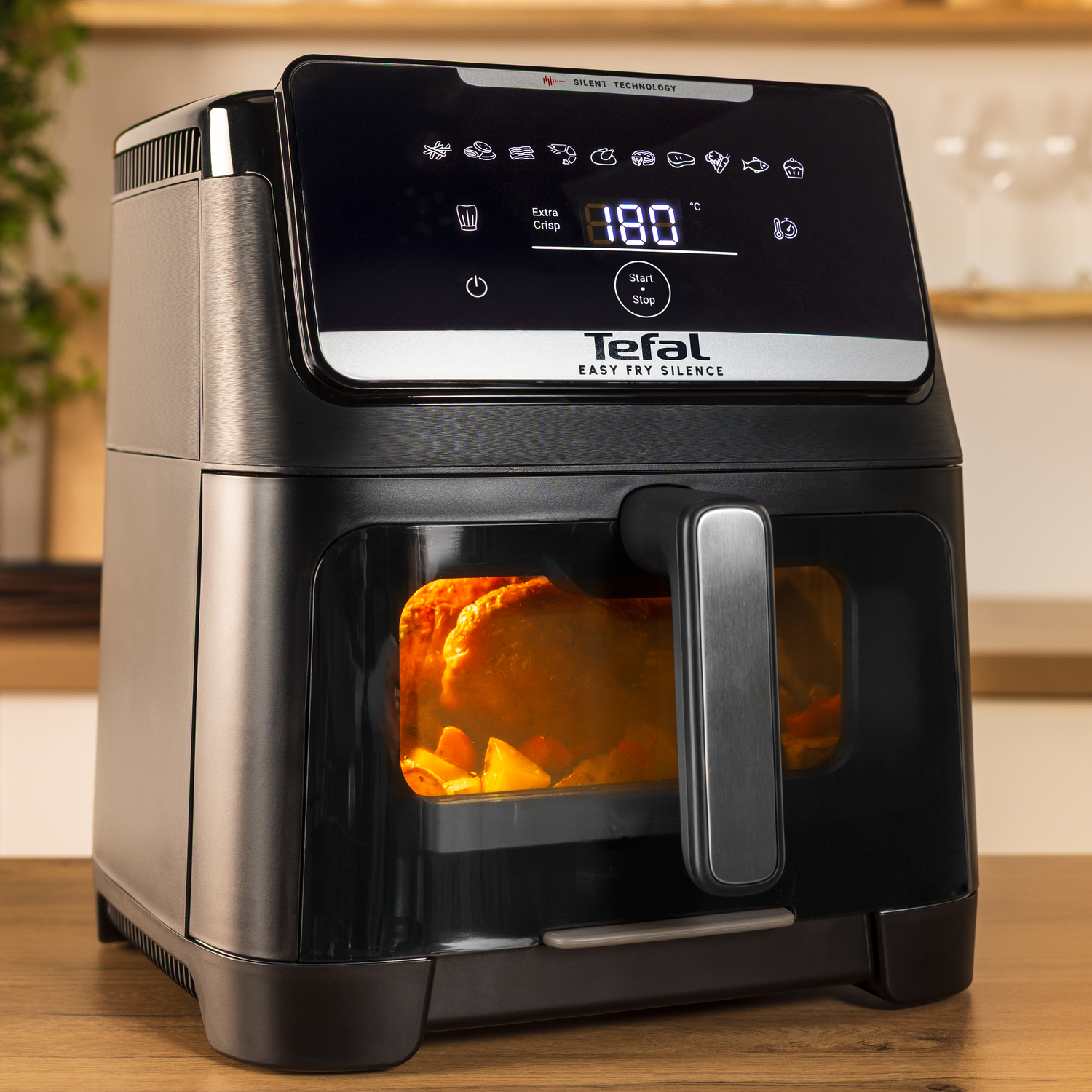 Black Tefal air fryer with food inside. Display shows 180 degrees. A wooden countertop and blurred background are visible.