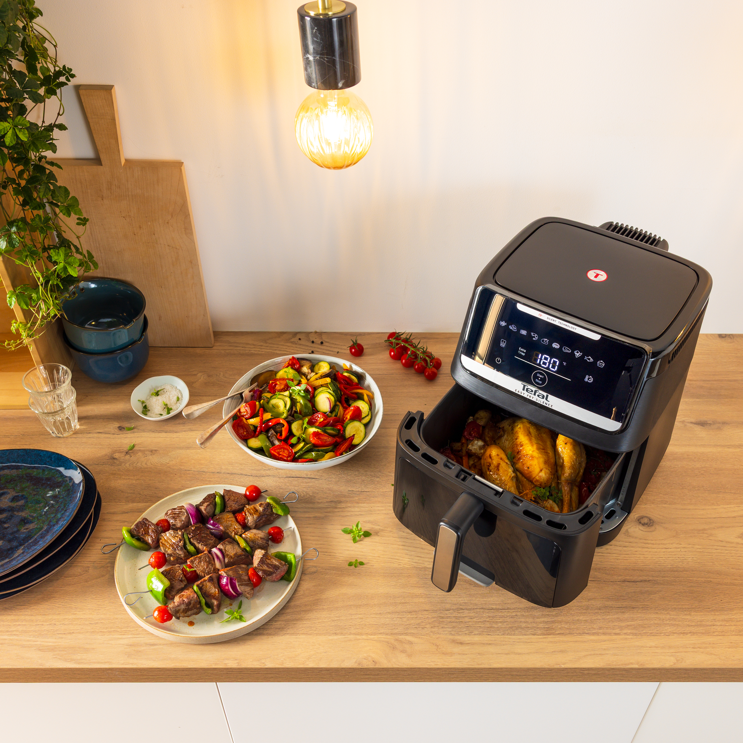 A black air fryer with chicken inside. Skewers with meat and vegetables, salad, and bowls on a wooden surface. A hanging light.