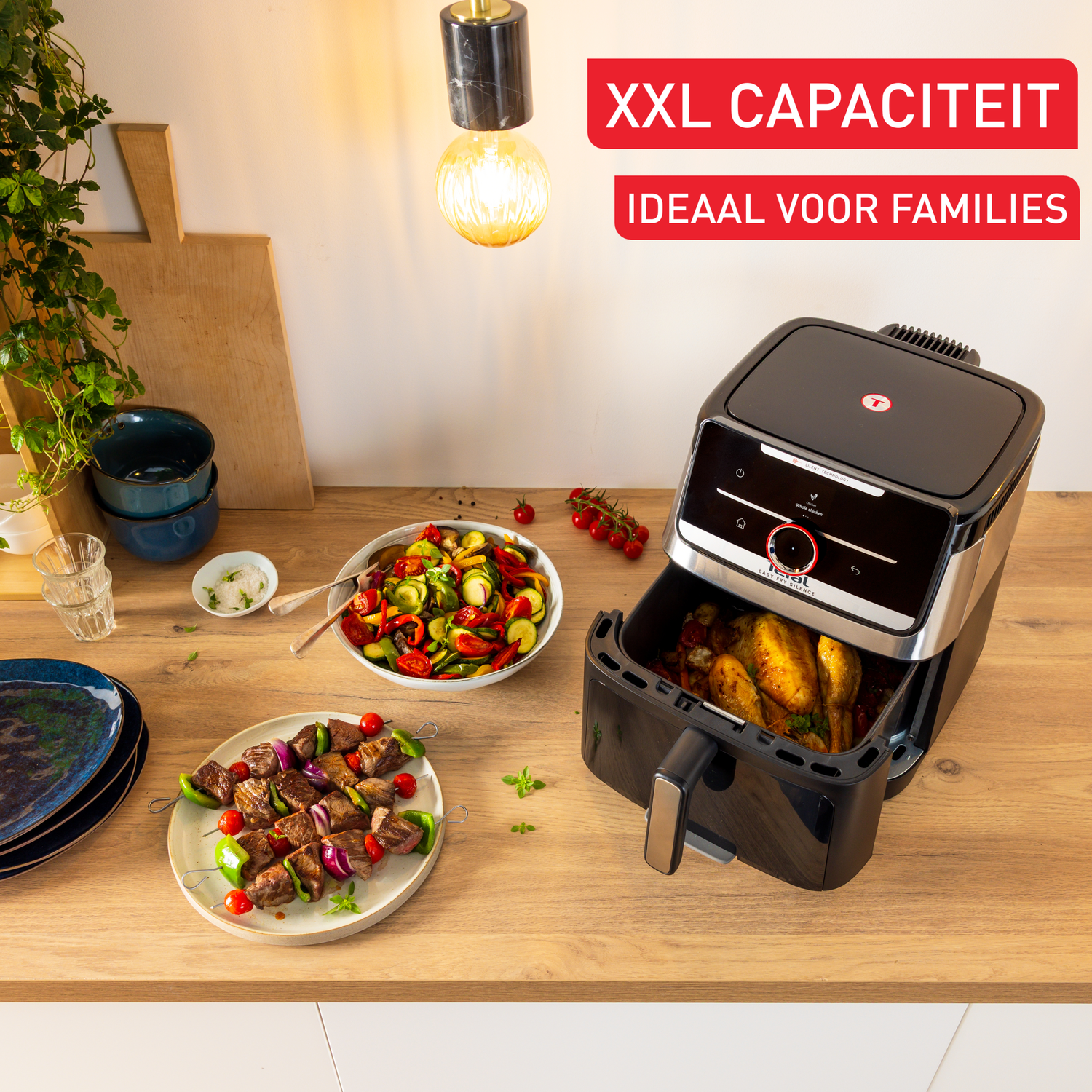 An air fryer with a chicken inside, with skewers and a vegetable salad on the table, and text that says 'XXL CAPACITEIT' and 'IDEAAL VOOR FAMILIES'.