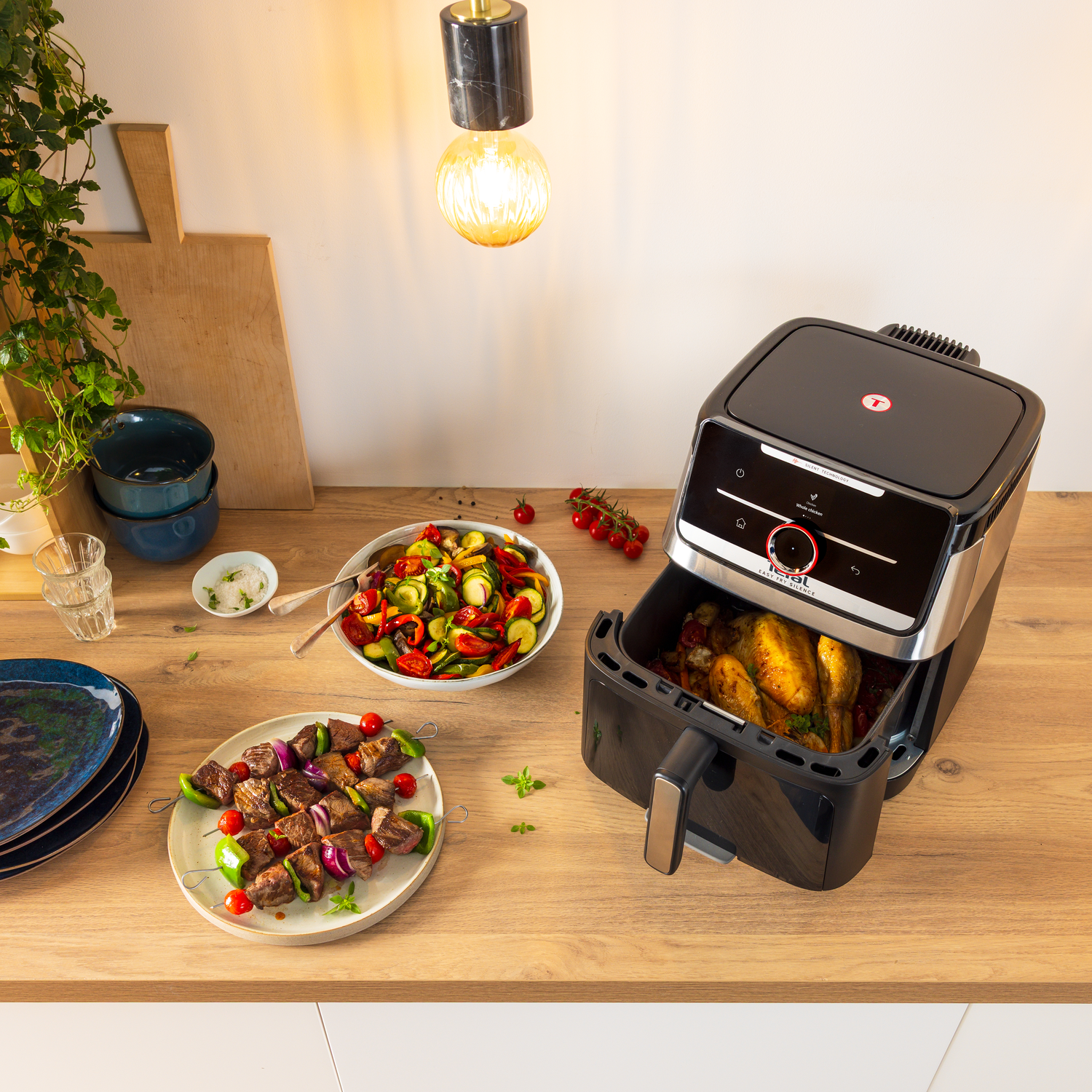An air fryer with a chicken inside, skewers, salad, and bowls on a wooden surface. A light hangs from the ceiling.