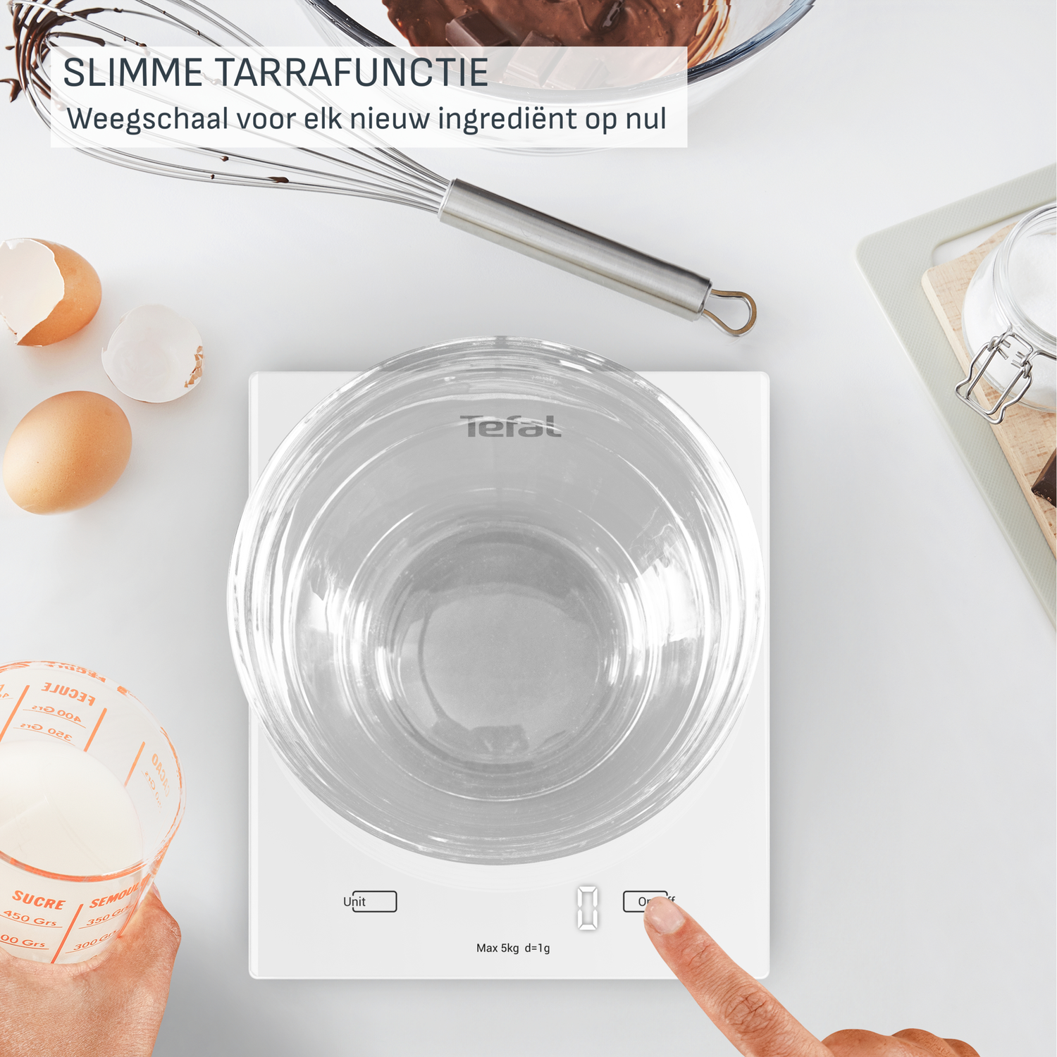 A kitchen scale with a bowl is on, next to it are eggs, a hand is pressing the button. Ingredients are around.