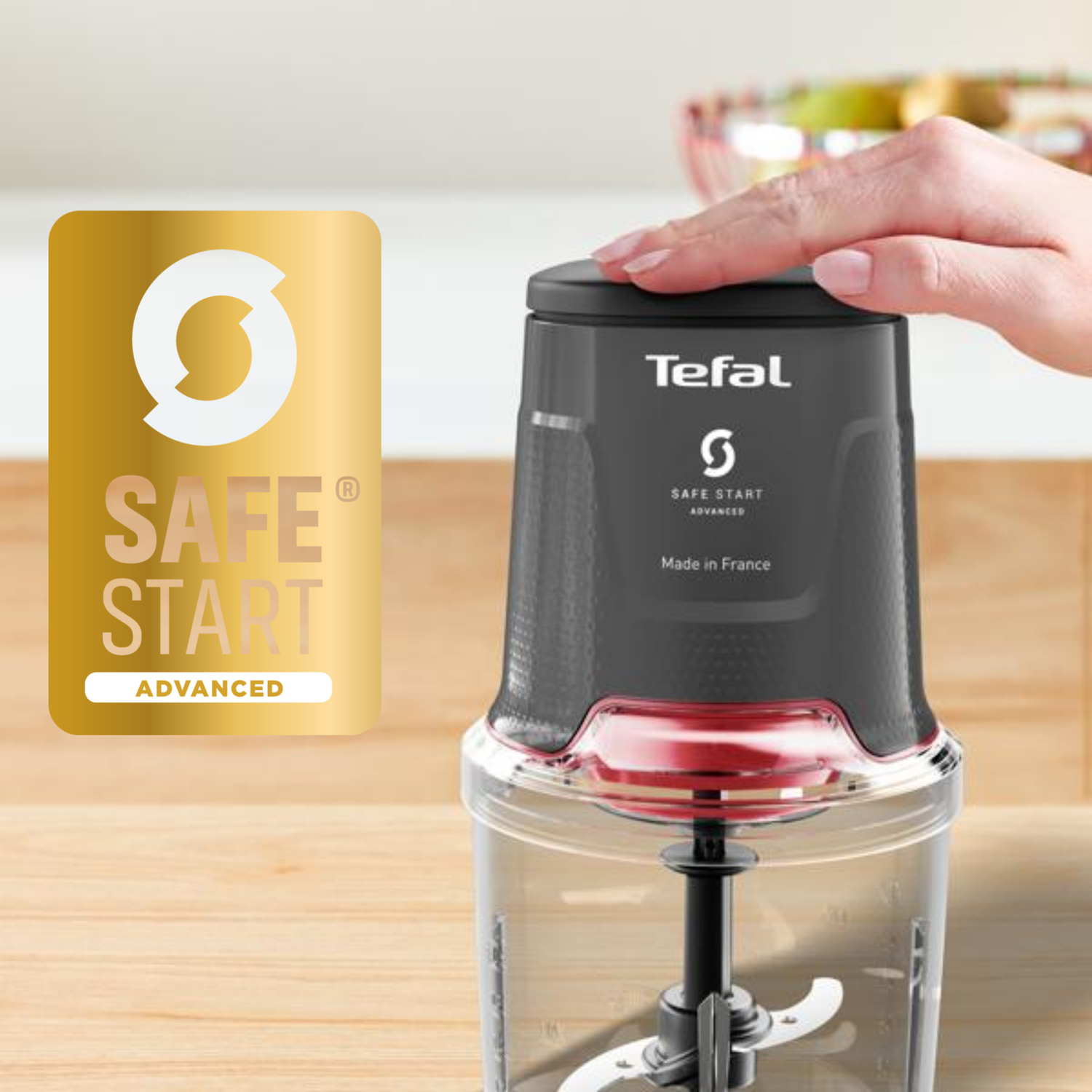 A hand presses on a black and red Tefal food chopper. A gold SAFE START logo is on the left. Chopper is on a wooden surface.
