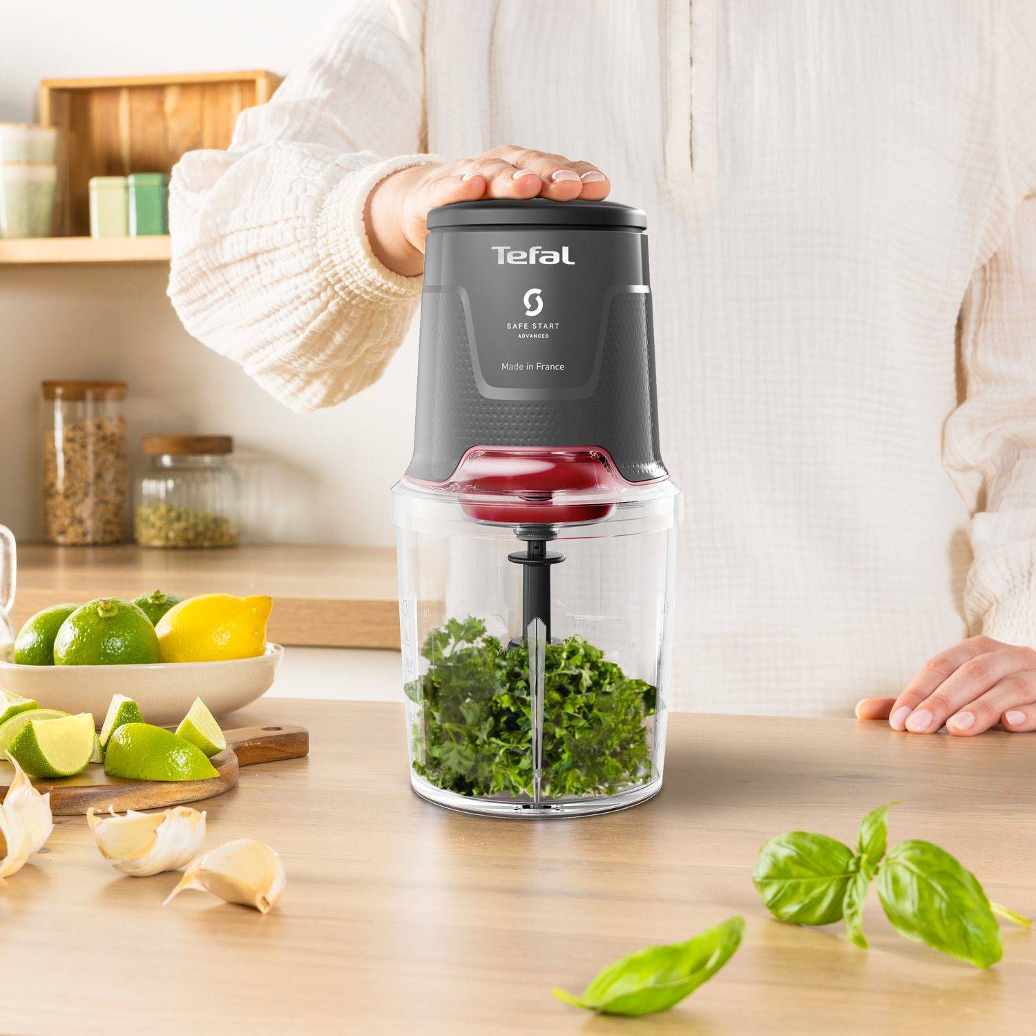 A person using a Tefal mini chopper. Limes, lemons, and garlic are on the wooden table. Basil leaves are also present.