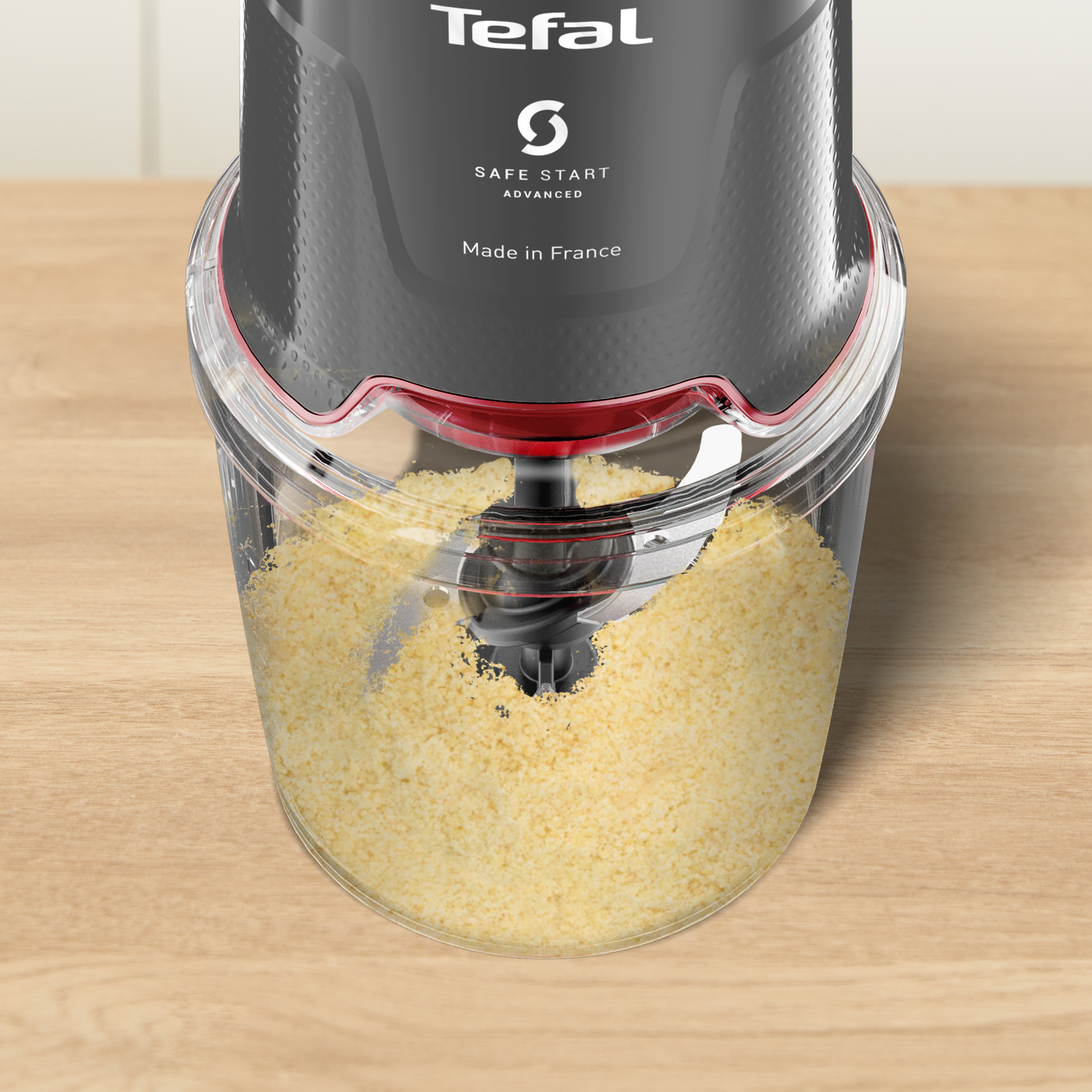 A Tefal food processor is filled with yellow food. The base is clear plastic, and the top is black with white text on a wooden surface.