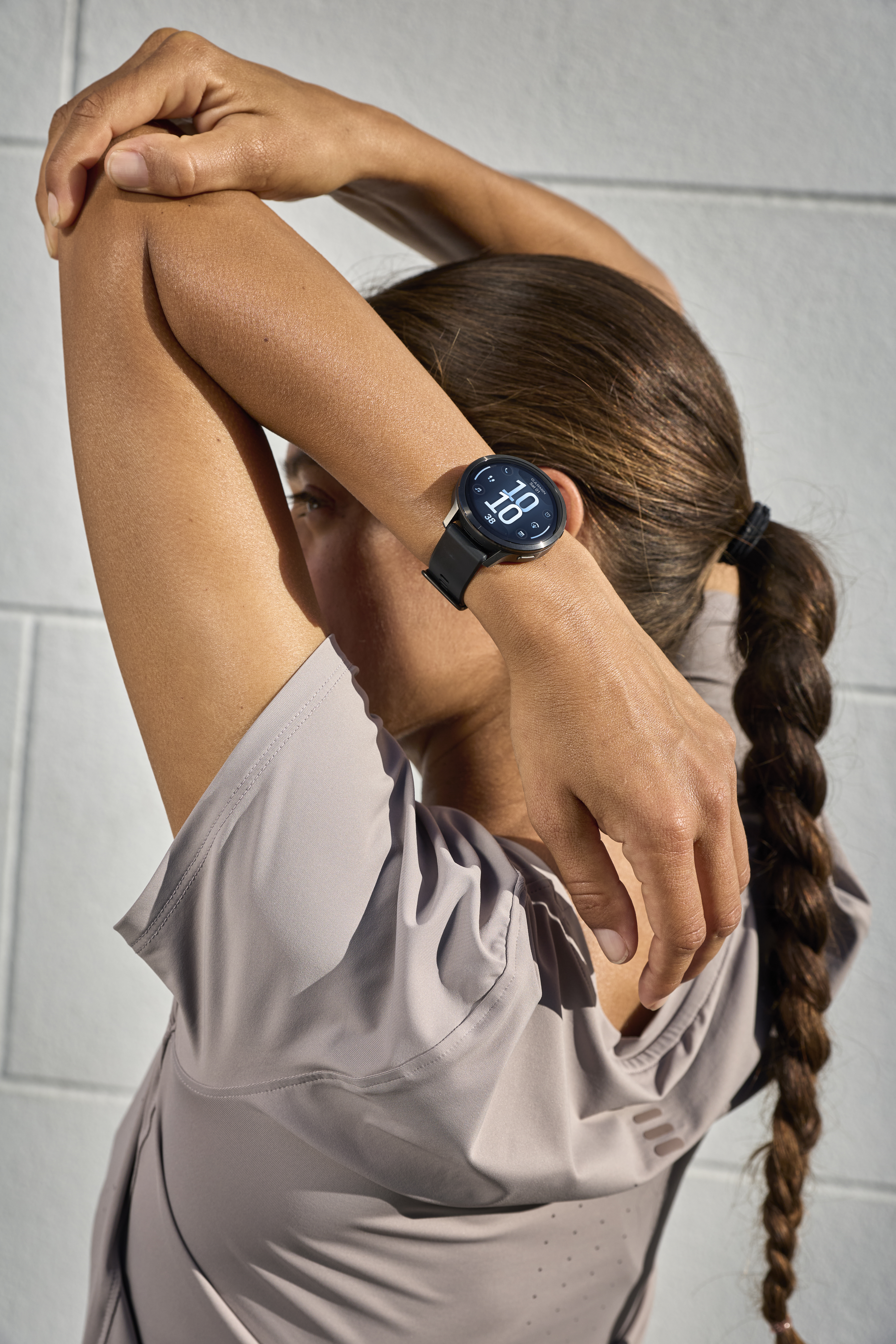 Eine Frau dehnt sich, Arm erhoben, mit Smartwatch. Sie trägt ein graues Sportoberteil. Hintergrund ist eine weiße Wand.