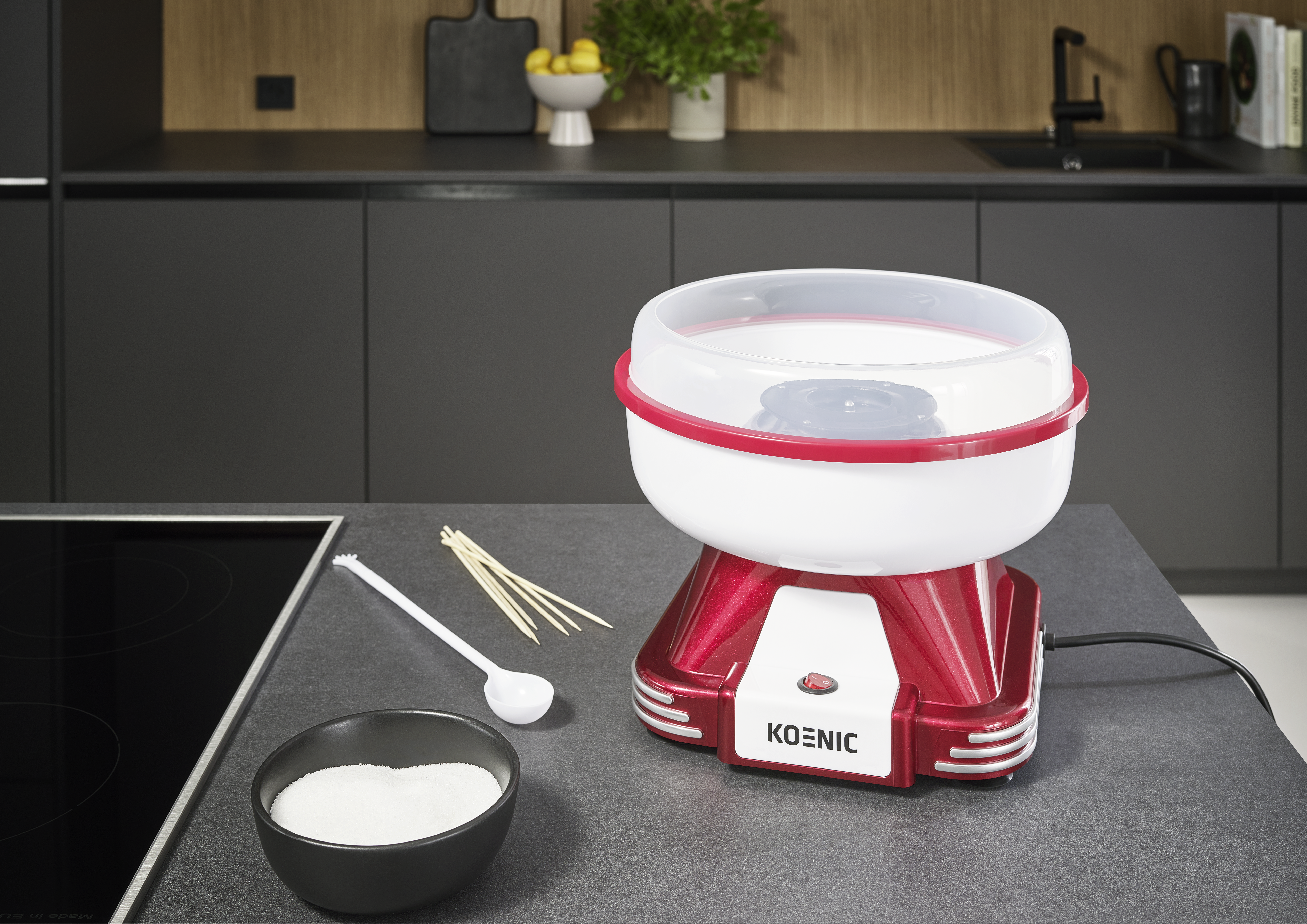 Ein rot-weißer Zuckerwattemaschine steht auf einer grauen Theke, mit Zucker, Stöcken und einem Löffel daneben. Küche im Hintergrund.