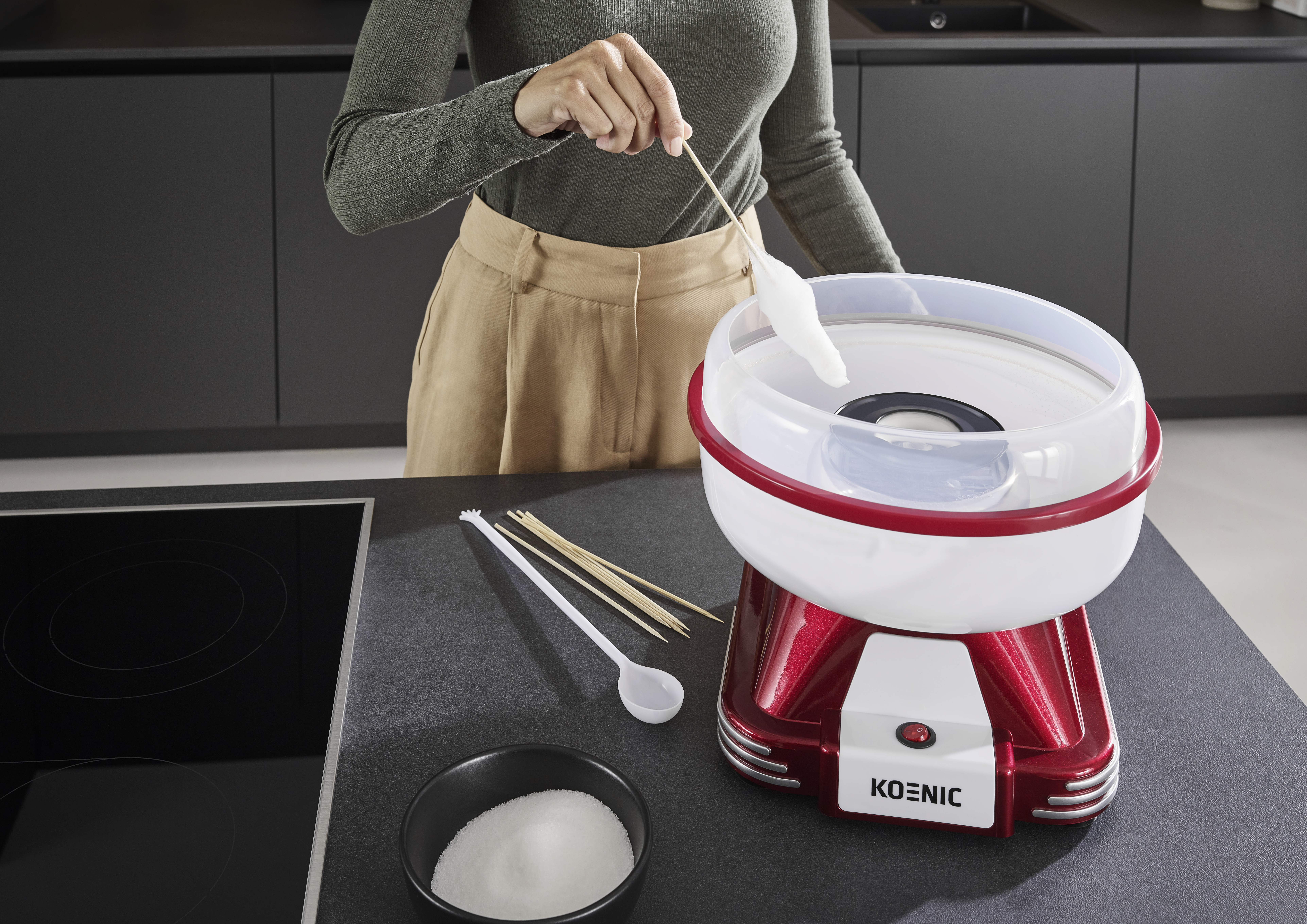 Eine Person stellt mit einer rot-weißen Maschine Zuckerwatte her. Zutaten und Werkzeuge liegen auf der Theke. Die Person trägt Grün und Beige.